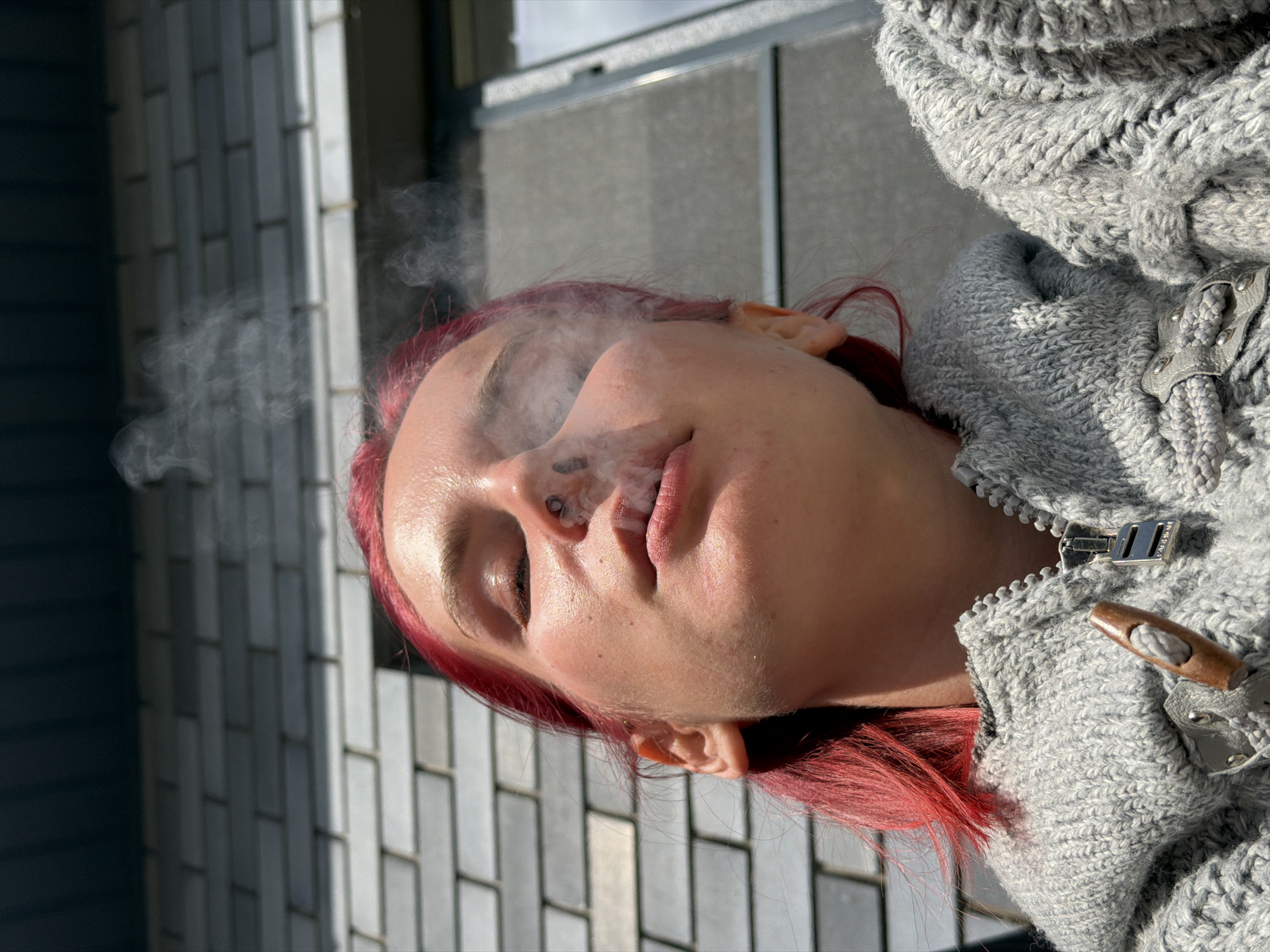 A low-angle, close-up shot shows a young woman with bright red hair, her eyes closed, exhaling a plume of smoke. She is wearing a thick, grey knit sweater with a toggle closure. The background is out of focus, but appears to be an exterior wall made of grey brick or stone, suggesting an outdoor urban environment. The lighting indicates it is daytime. There is no information to suggest the location is Chaikovsky, Russia.