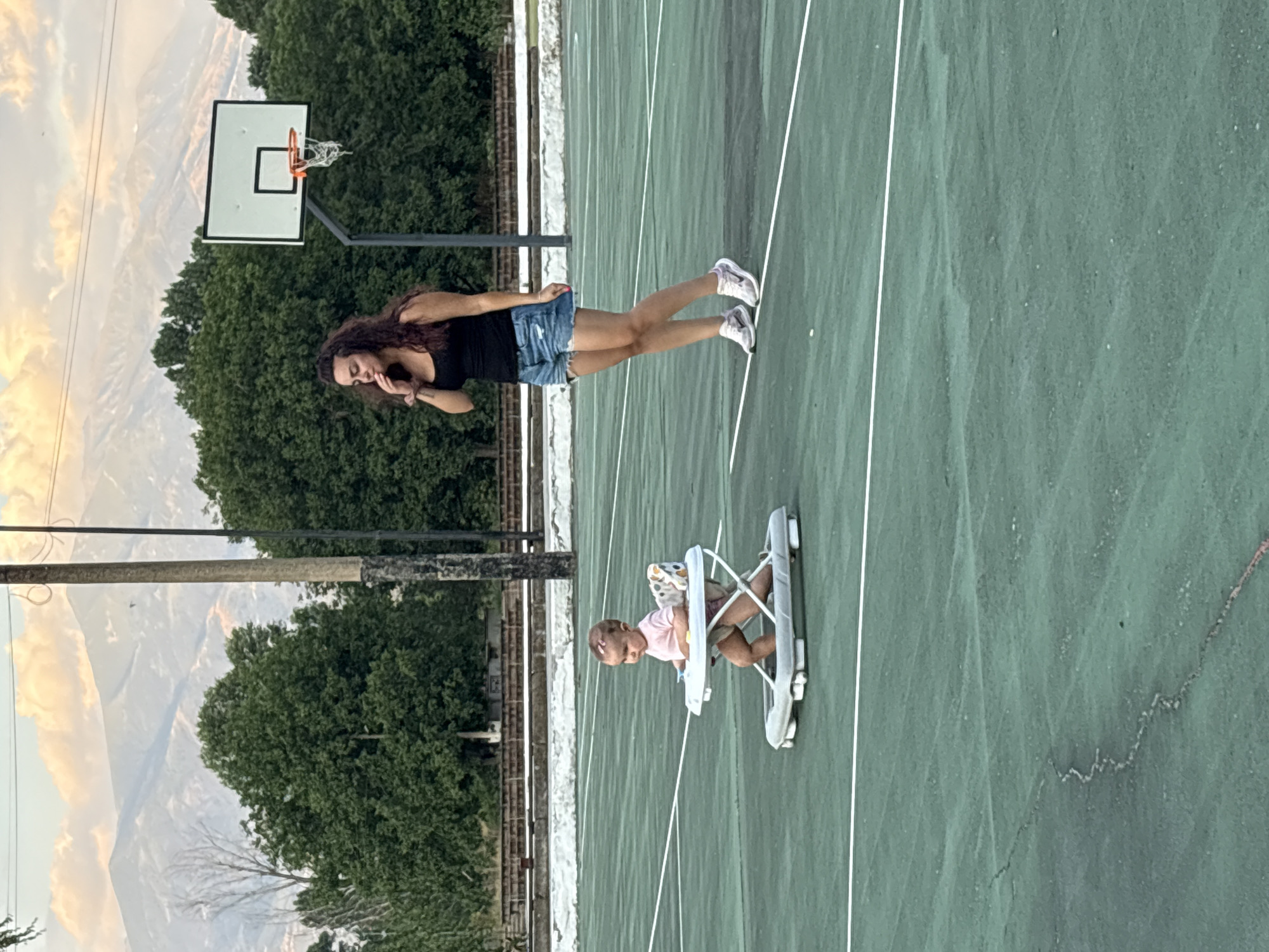 The image shows a young child in a baby walker on a green tennis court. A woman stands behind the child, looking down and to the side. In the background, there are trees, a basketball hoop, and mountains. The location is identified as Boboshevo, Bulgaria. The scene appears to be outdoors during the day, with a natural light. The woman is wearing a black tank top and denim shorts, and the child is wearing a pink outfit.