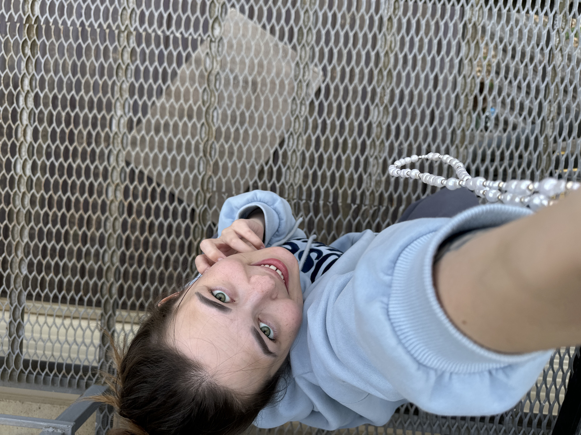 A young adult female is taking a selfie while standing on a metal grate platform. She is wearing a light blue hooded sweatshirt and dark pants. She is holding a string of white pearls in her right hand. The location appears to be an industrial or urban area, suggested by the metal grate flooring and a building visible in the background. The image perspective is from above, looking down at the subject. It is not possible to confirm the location as Ilyinsky, Russia based solely on the visual information provided.