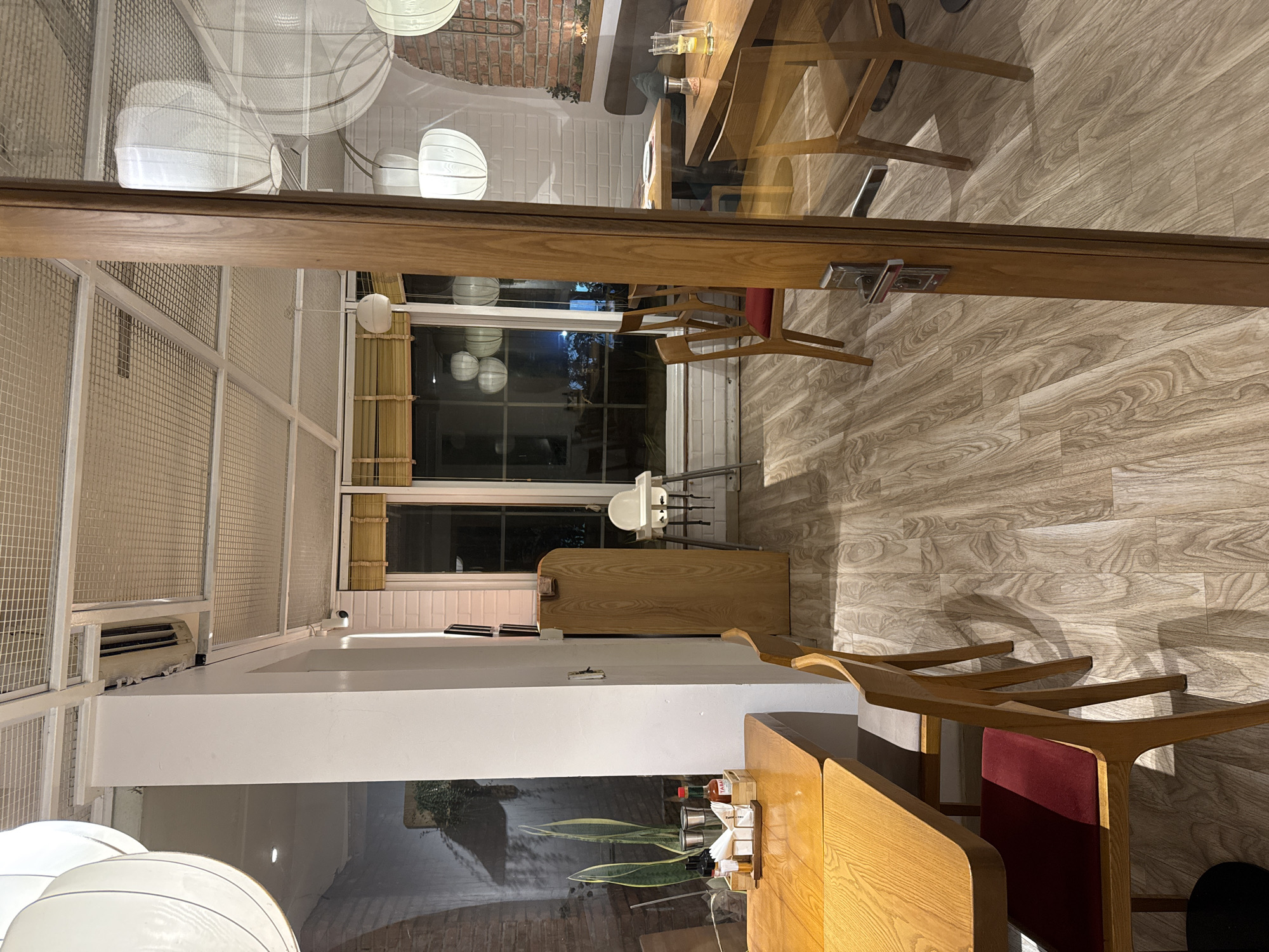 The image displays the interior of a restaurant with a modern, minimalist design. The scene is viewed through a glass door, which reflects some of the interior elements. Inside, several wooden tables and chairs are arranged, with a focus on clean lines and natural wood tones. One table in the foreground is set with condiments, napkins, and what appears to be a glass of drink. In the background, more tables and chairs are visible, along with large white pendant lights hanging from the ceiling.
The setting appears to be a contemporary dining establishment, possibly in Hanoi, Vietnam, as indicated by the context. The lighting suggests it could be daytime or early evening, with artificial lights supplementing the natural light. The overall atmosphere is clean, organized, and inviting, with the wooden elements and ample lighting contributing to a warm and sophisticated ambiance. There are no people visible in the image, emphasizing the architectural and design aspects of the space. The floors are wooden, with a subtle wood-grain pattern. The walls in the background are a mix of white subway tiles and exposed brick, adding texture and visual interest. The large windows or glass partitions allow light to filter through, creating soft shadows on the floor. The reflection on the glass door shows a slightly distorted view of the interior, adding depth to the image.