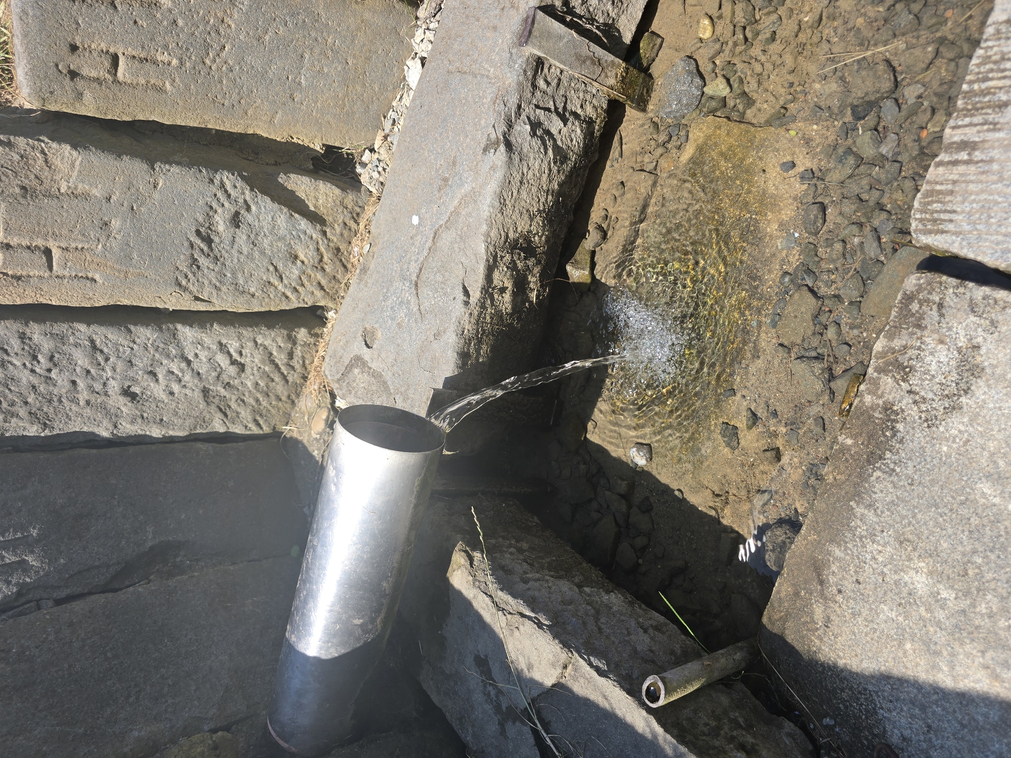 A close-up, high-angle shot shows a stainless steel pipe delivering water from a large stone structure into a shallow pool of water. The water from the pipe creates ripples on the surface of the pool. The surrounding environment consists of large, rough-hewn stone blocks, some of which are inscribed with simple markings. The pool bed is composed of sand and small pebbles, with a few blades of grass visible. The lighting suggests a sunny day, casting shadows on the stone surfaces. The image appears to be of a water feature, possibly a natural spring, located in Pustiměř, Czechia.