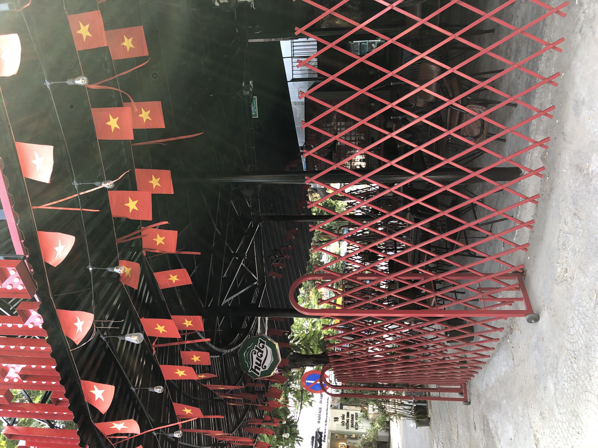 The image depicts the exterior of an establishment in Da Nang, Vietnam. A red, expandable metal barrier with diamond-shaped openings is positioned in the foreground, extending across the width of the frame. This barrier is on wheels, suggesting mobility. Behind the barrier, an outdoor seating area is visible, with several tables and chairs, some of which appear to be made of wood and leather.
Overhead, a black corrugated metal roof is supported by dark metal poles. Strands of small red flags with yellow stars, resembling Vietnamese flags, are strung across the underside of the roof, interspersed with exposed light bulbs. A hexagonal green sign with white lettering, a logo or brand name, is suspended from the roof. Further back, an illuminated green exit sign with arrows is visible. The ground is paved with concrete, and the overall setting suggests a commercial area or restaurant with an outdoor component, decorated with nationalistic symbols.