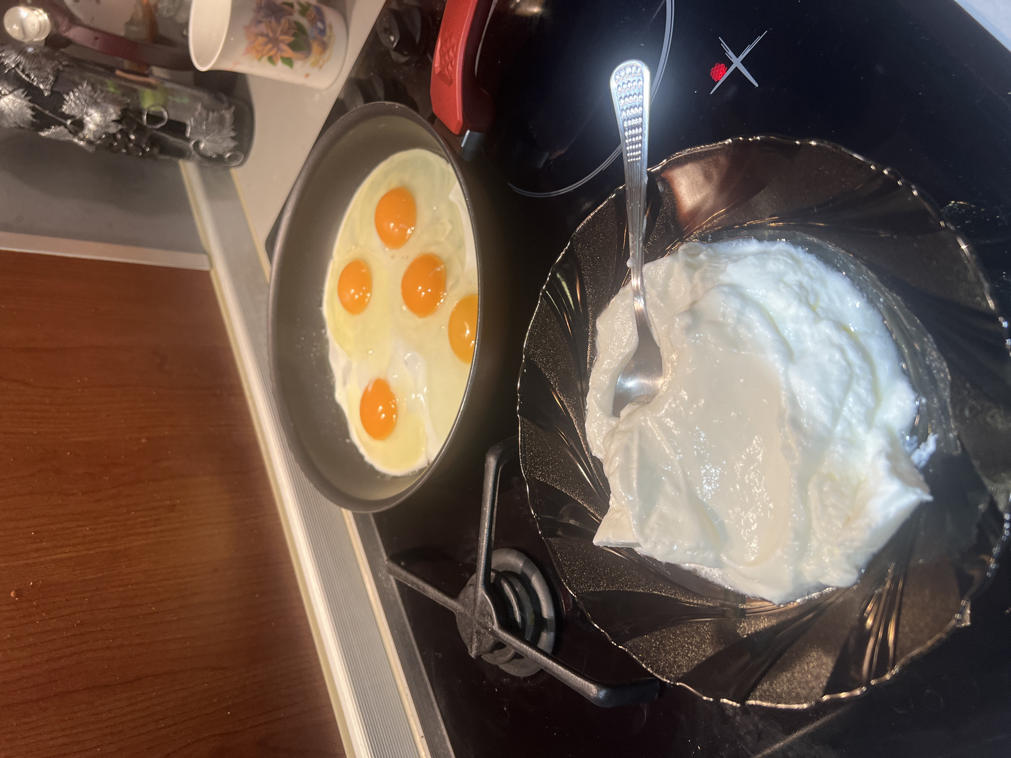 The image shows a still life composition of breakfast foods on a kitchen stovetop. A black frying pan contains six fried eggs with runny yolks. Next to the pan is a clear decorative bowl filled with a large dollop of white cream, likely yogurt or sour cream, and a silver spoon rests inside. The stovetop is black, with an induction coil and burner visible. The location is not discernible as Burgas, Bulgaria, from the image. The rest of the kitchen is partially visible on the left, with countertop items and a cabinet.