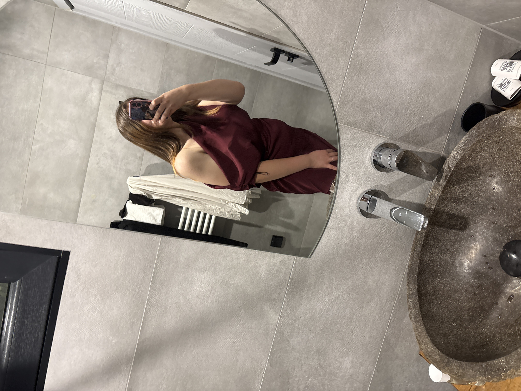 The image depicts a person taking a mirror selfie in a modern bathroom. The bathroom features large grey tiles on the walls and a dark grey countertop. A stone sink with chrome faucets is visible in the foreground. The person is wearing a maroon off-the-shoulder dress and is holding a phone to take the picture. In the reflection, a towel rack with white towels and a black garment is visible. The overall aesthetic suggests a contemporary and minimalist design. This scene is located in Dolno Cherkovishte, Bulgaria.