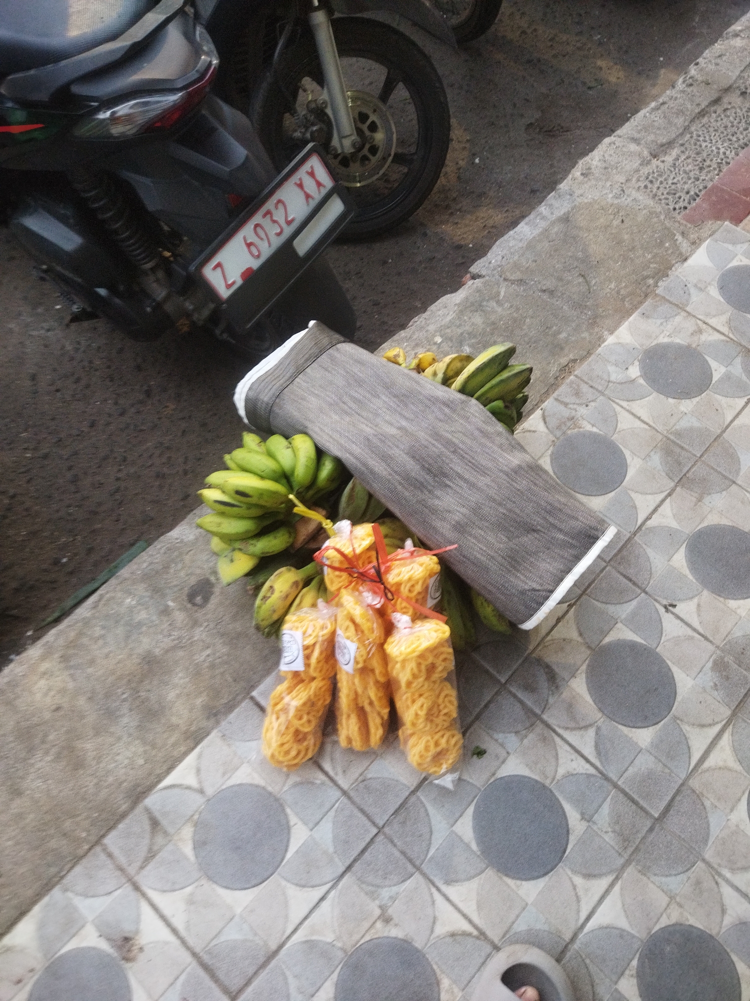 An overhead, slightly angled medium shot shows a motorcycle parked on a sidewalk in Banjar, Indonesia. The motorcycle's license plate reads "Z 6932 XX". Next to the motorcycle, a bunch of green bananas is laid out with several bags of yellow, coiled snacks tied with red string. The sidewalk is made of grey and white patterned tiles, and the curb is rough concrete. A dark asphalt road is visible behind the motorcycle.