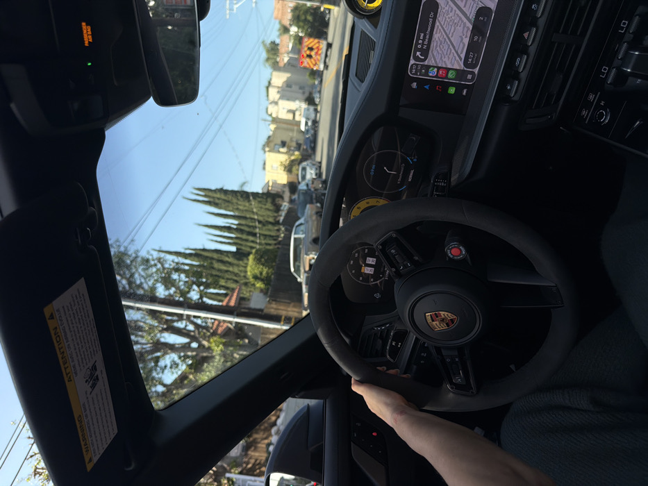 An eye-level shot from inside a Porsche captures the driver's perspective. The black steering wheel with a Porsche logo is prominently featured, alongside the dashboard displaying navigation and speed. Outside the window, tall trees and buildings line the street under a bright sky. A sun visor with a warning label is visible in the upper left corner. The passenger airbag indicator is also visible. Overall, the image conveys a sense of driving through a residential area in a luxury vehicle.