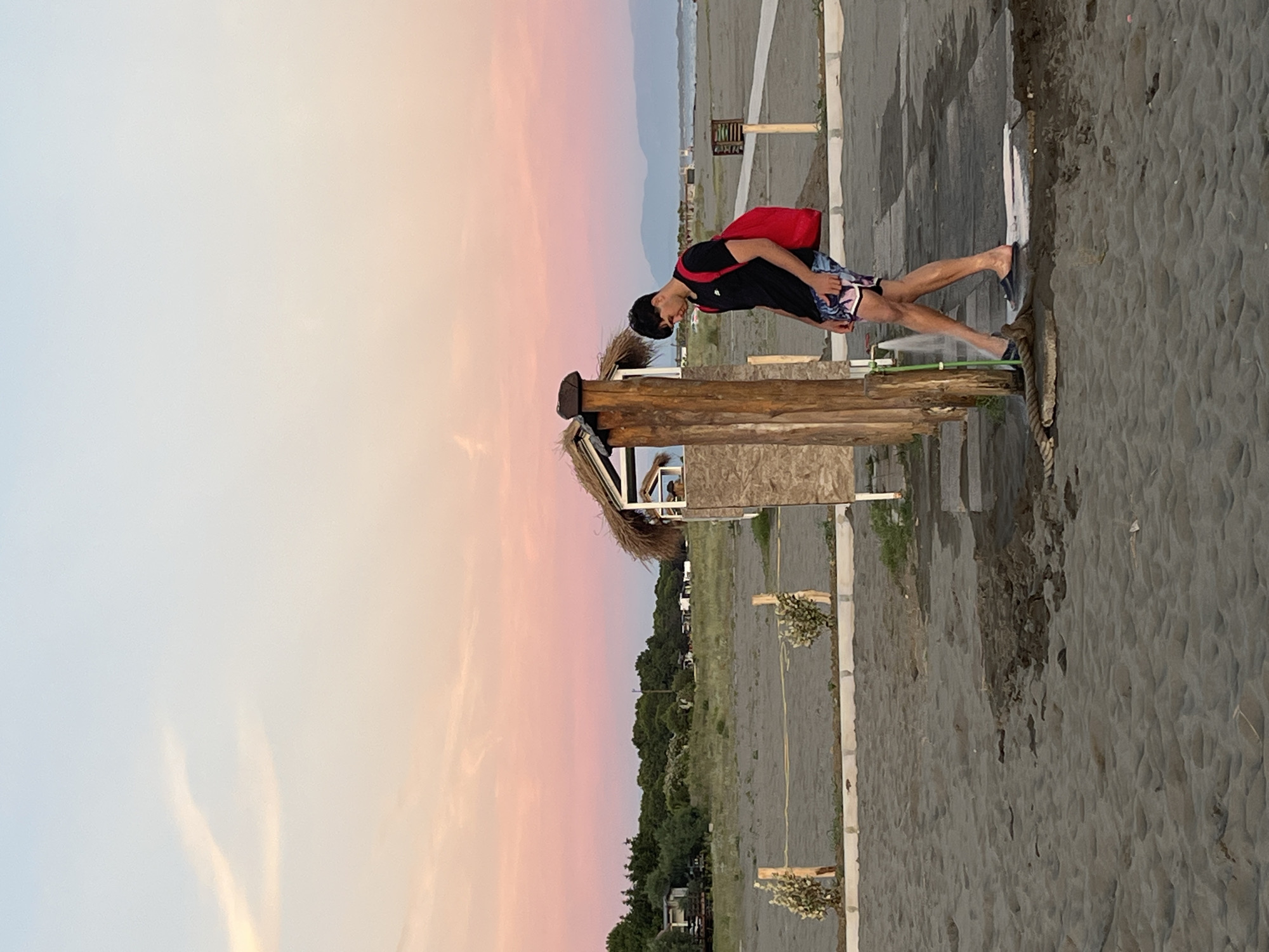 A young man with dark hair is standing on a sandy beach, rinsing his feet under a shower. He is wearing a black t-shirt and patterned shorts, with a bright red backpack slung over his left shoulder. The shower is attached to a wooden post, and water is spraying onto the sandy ground, creating a small puddle. In the background, there is a small, thatched-roof structure that appears to be a changing room or restroom. Further back, a line of trees separates the beach from a low-lying area with some buildings and vehicles. The sky above is a gradient of soft pinks and blues, indicative of a sunset or sunrise. The location is likely near the city of Ulcinj, Montenegro, which is close to Pistula.