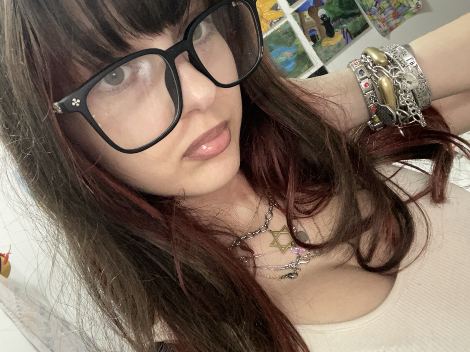 A woman with dark hair and bangs, wearing black-framed glasses, looks at the camera. She has layered necklaces, one with a Star of David pendant. Her arm is adorned with multiple bracelets. She wears a cream-colored top. There are various framed artworks visible in the background. Her hair has red highlights.