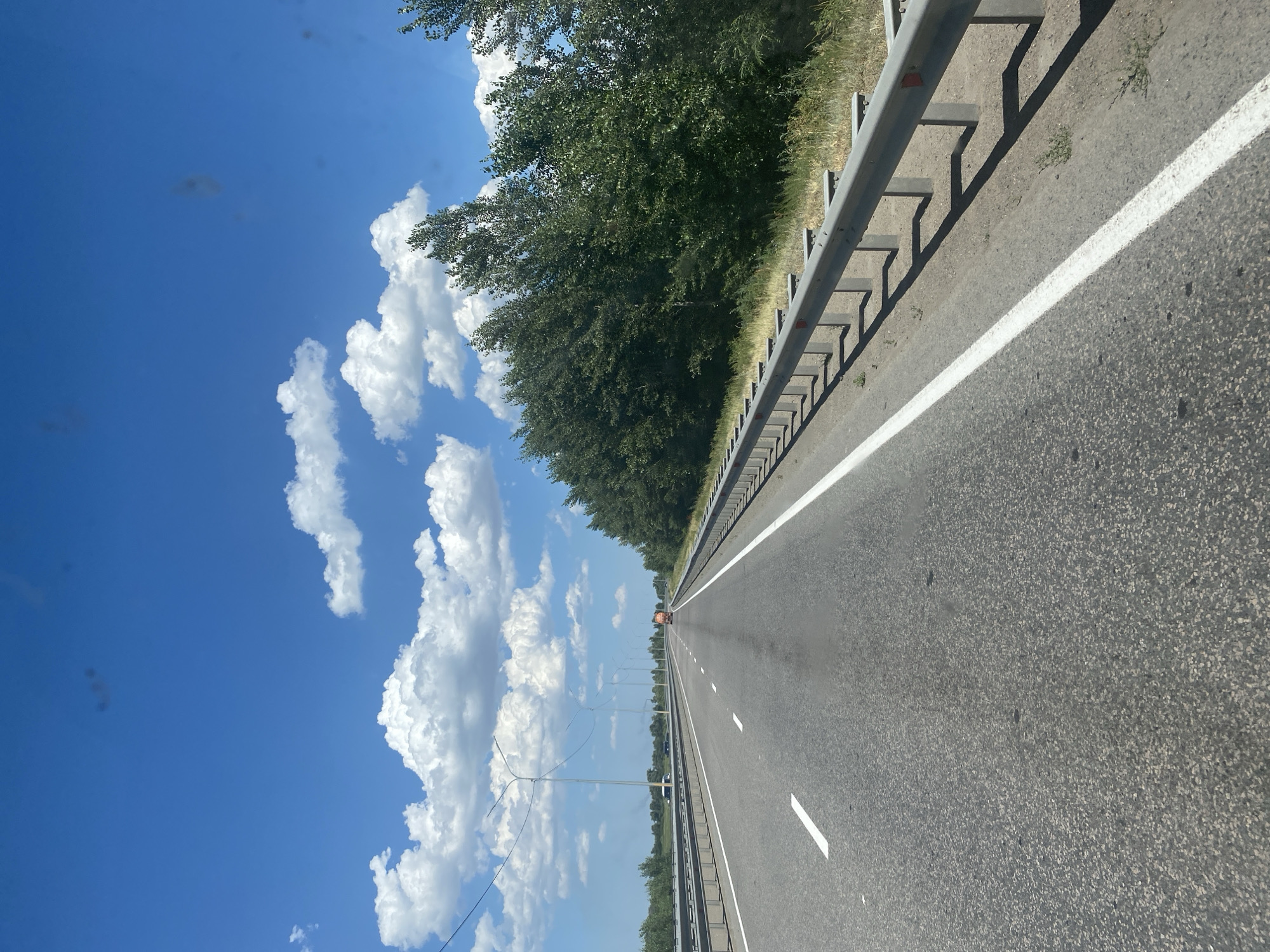 The media file depicts a highway scene on a clear day with a bright blue sky and fluffy white clouds. The road is asphalt, marked with white lines, and has a metal guardrail on the right side. A dense line of green trees borders the highway on the left. In the distance, a few cars and a bright orange vehicle, possibly a construction or maintenance truck, can be seen on the road. Tall lampposts with dangling wires line the side of the highway. The sun appears to be high in the sky, suggesting it is daytime. The overall impression is one of a travel or transit scene. There is no visible text in the image. The location context provided is "Aqsu city administration, Kazakhstan".