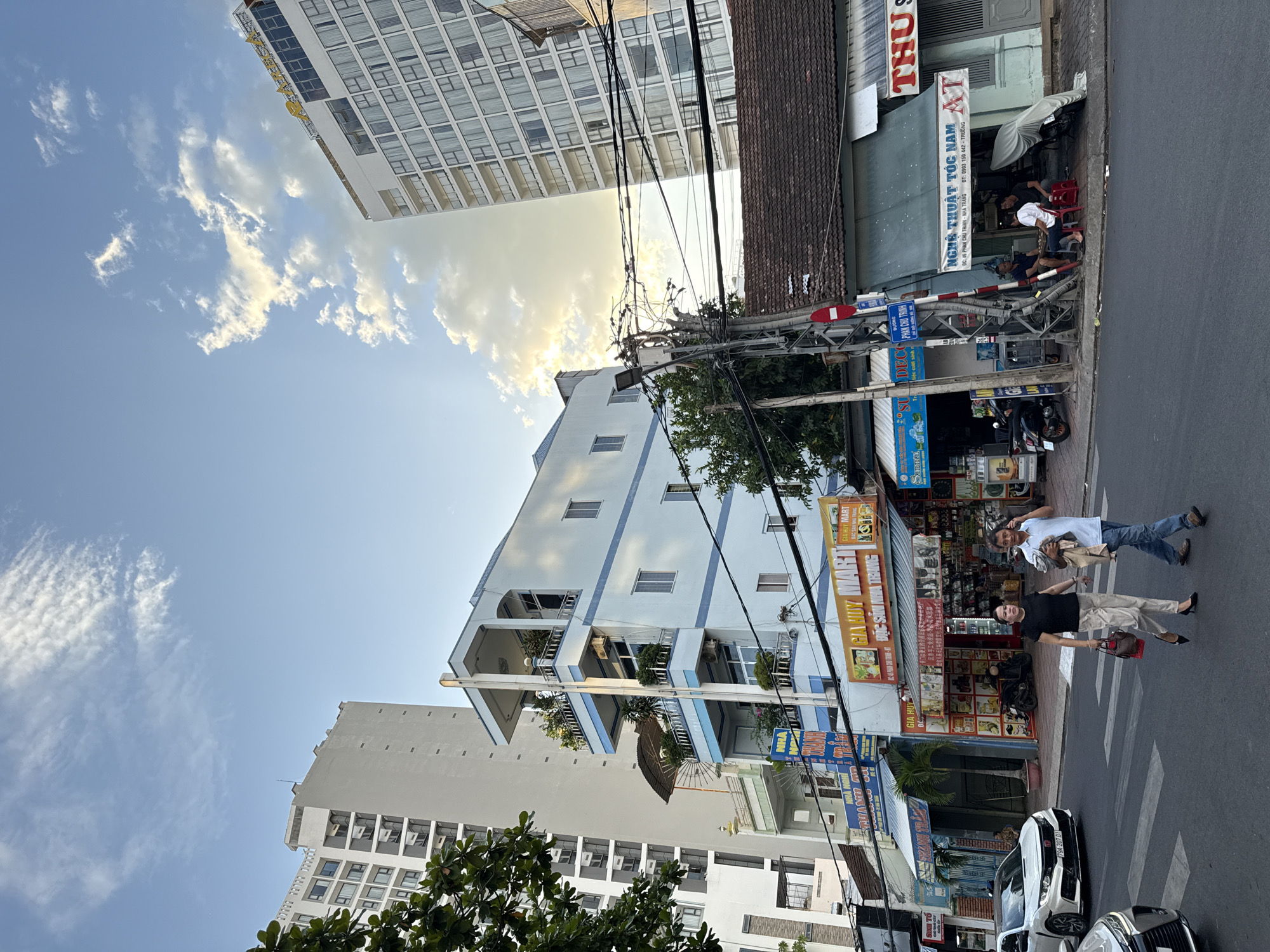 The image depicts a street scene in Nha Trang, Vietnam, under a sky with scattered white clouds illuminated by the sun. In the foreground, two individuals, a woman and a man, are walking across a street. The woman, wearing a black top and light-colored pants, carries a red handbag. The man, dressed in a light blue shirt and jeans, carries a folded garment. They are crossing a pedestrian crossing.
Several buildings line the street. To the left, a tall, modern white building with many windows stands prominently. Adjacent to it is a lighter blue, multi-story building with balconies adorned with plants. Further down the street, to the right, a row of older, single-story shops with tiled roofs are visible. One shop has a sign that reads "GIA HUY MART" with text in Vietnamese. Another sign reads "THU S". A traffic light pole with numerous wires is situated on the sidewalk.
In front of one of the older buildings, three men are seated on red plastic chairs, seemingly engaged in conversation or a game. A covered motorcycle is parked nearby. Cars are also present on the street, including a white sedan on the left side of the frame. The overall atmosphere suggests a typical urban environment during daylight hours.