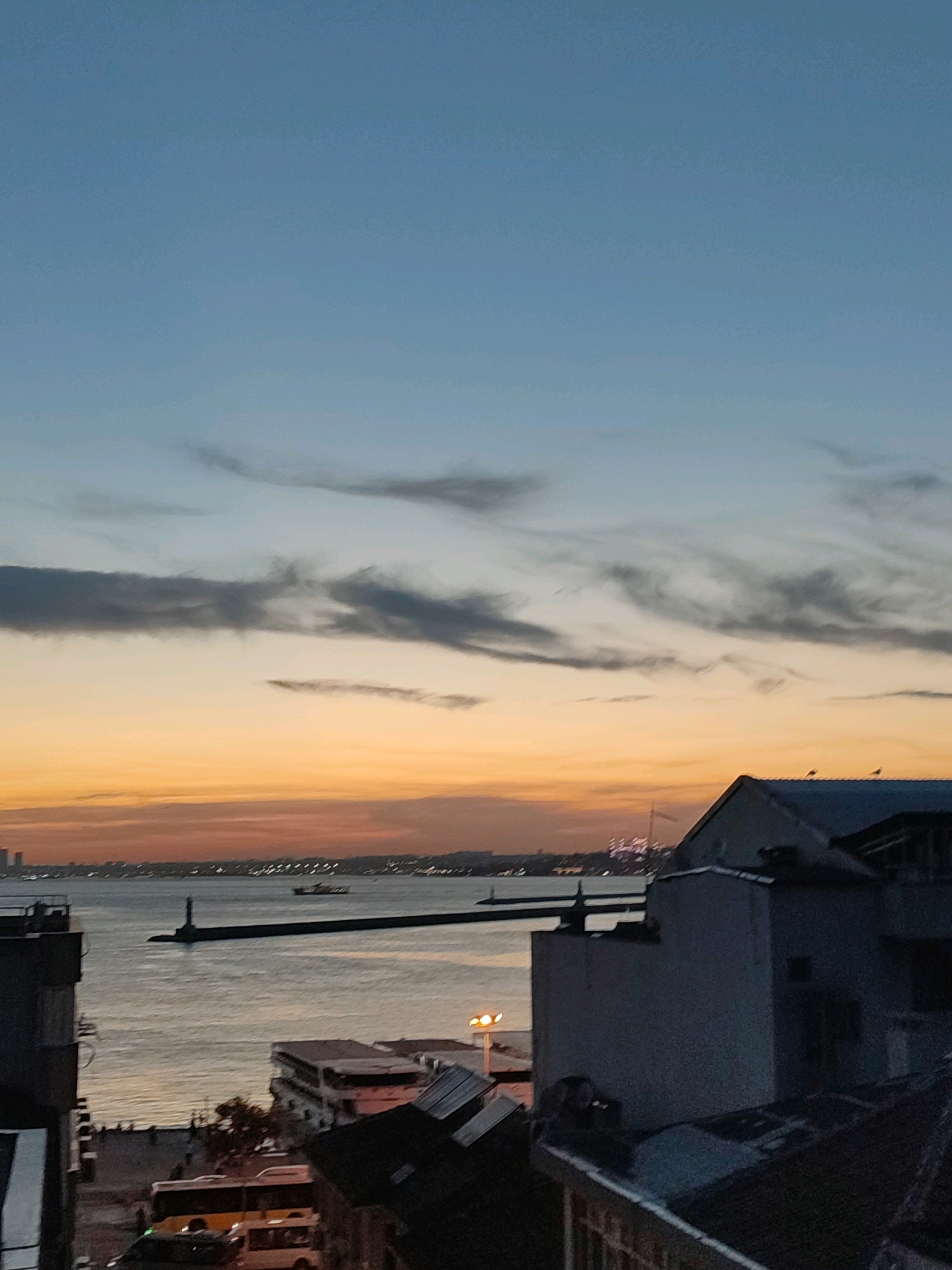 This is a vertical, medium-shot photograph of a dusk scene in Kadıköy, Turkey. The sky is a gradient of deep blue at the top, transitioning to lighter blues and then to a band of orange and pink hues near the horizon, indicating sunset. Wispy grey clouds are scattered across the sky.
In the foreground, the rooftops of buildings are visible, some appearing dark and silhouetted. On one roof, three small dark shapes, likely birds, are perched. A street scene is partially visible below the rooftops, with a yellow bus and a white van parked. Pedestrians are present on what appears to be a walkway.
The middle ground features a body of water, likely the Sea of Marmara, with calm surface reflecting the sky's colors. A long, dark pier or breakwater extends into the water, with a lighthouse at its end. A ship is visible in the distance on the water.
The background shows a distant cityscape with buildings of varying heights, some of which are illuminated, creating a glow against the darkening sky. The overall impression is of an urban coastal setting at twilight.