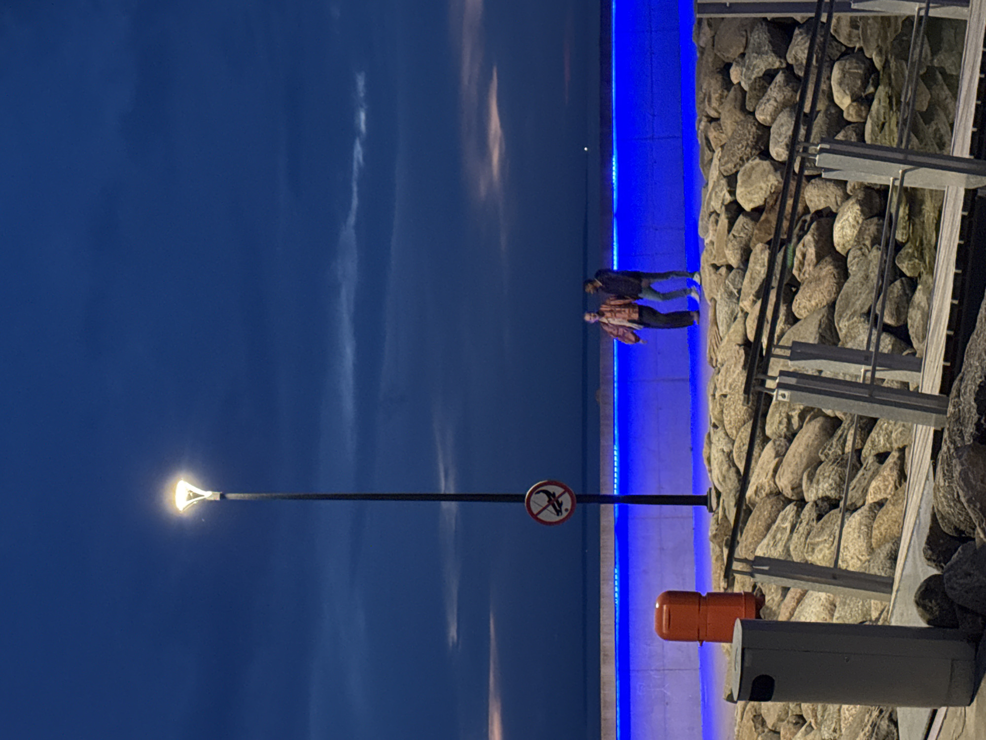 The image depicts a waterfront scene at dusk or night, located in Tallinn, Estonia. The sky is a dark, deep blue with scattered, darker clouds. A tall, dark streetlamp with a bright, warm-toned conical LED fixture illuminates the upper portion of the frame. Attached to the streetlamp pole, at approximately half its height, is a circular "no fishing" sign, featuring a red border, a white background, a red diagonal line, and a black stylized fish on a hook.
Below the streetlamp, an orange cylindrical public waste bin is positioned to the left. A long concrete wall, illuminated along its top edge by a continuous strip of bright blue LED lighting, runs horizontally across the mid-ground. Behind this wall, two individuals stand on a paved surface. The person on the left wears a patterned jacket with shades of brown and possibly red, dark trousers, and dark shoes, facing partially towards the other person. The person on the right wears a dark jacket, blue jeans, and light-colored shoes, also oriented towards the first individual, suggesting interaction.
The area in front of the concrete wall consists of a lower section filled with numerous large, irregularly shaped grey and brown rocks. Extending from the bottom-right towards the center is a wooden walkway or ramp with light grey metal railings, descending into the rocky area. In the bottom-left corner, a grey cylindrical public waste bin with a black opening is partially visible. Beyond the blue-lit wall, a flat, dark body of water meets the horizon beneath the twilight sky.