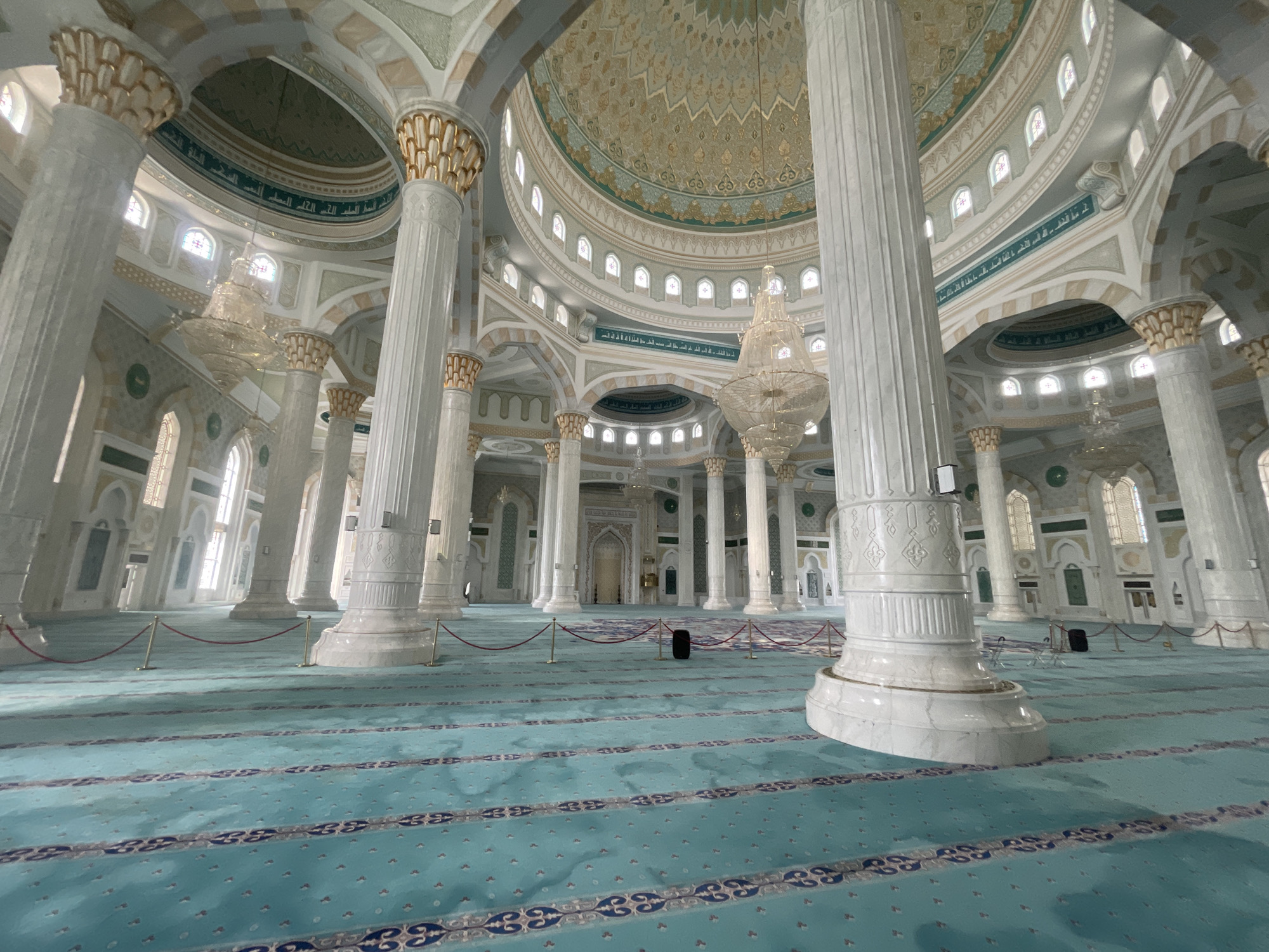 The image depicts the interior of the Hazrat Sultan Mosque in Astana, Kazakhstan. The mosque features a vast prayer hall adorned with numerous white marble columns, some with gilded capitals, supporting a series of domes. Above, intricate geometric patterns decorate the main dome, and ornate chandeliers hang from the ceiling. Light enters through windows along the walls. A turquoise carpet with patterned borders covers the floor, marked with rows for prayer. Red ropes and gold stanchions delineate areas within the hall. No people are visible in the main part of the hall.