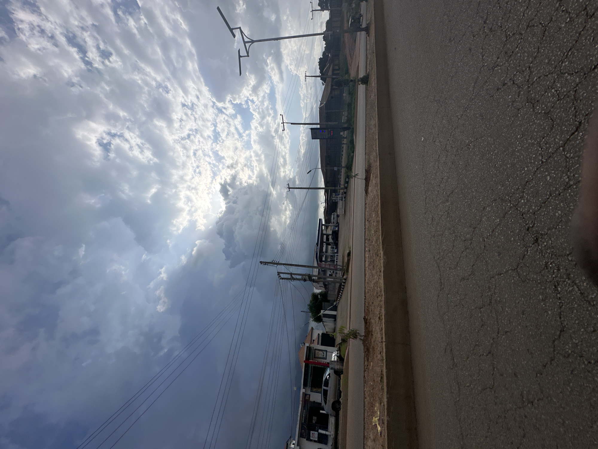 This media file depicts a roadside scene in Bukuru, Nigeria, with a focus on the sky and the surrounding infrastructure. The sky is dramatically cloudy, with sunlight breaking through in some areas, suggesting a dynamic weather pattern, possibly a storm approaching or receding. The clouds are a mix of bright white and darker grays, creating a sense of depth and movement.
In the foreground, a paved road with visible cracks is on the right side of the image. To the left of the road, there's a concrete curb and then a sidewalk area. Beyond the sidewalk, a series of utility poles with multiple wires stretches along the scene. These poles are characteristic of urban or semi-urban environments where electricity and telecommunication lines are overhead.
Several buildings and structures are visible behind the utility poles. These appear to be low-rise buildings, some with storefronts. There are indications of commercial activity, such as signage and what might be a gas station in the distance. A few vehicles, including what looks like a white van or truck and a smaller car, are parked or driving along the road. The overall impression is of a typical street scene in a populated area.
The time of day is likely late afternoon or early evening, given the quality of light and the dramatic cloud formations. There are no people clearly visible in the frame, so the activity is primarily ambient, representing the infrastructure and natural elements of the location. The emotional tone is somewhat dramatic due to the sky, but also mundane, depicting everyday life. No specific text is legible due to image quality and distance.