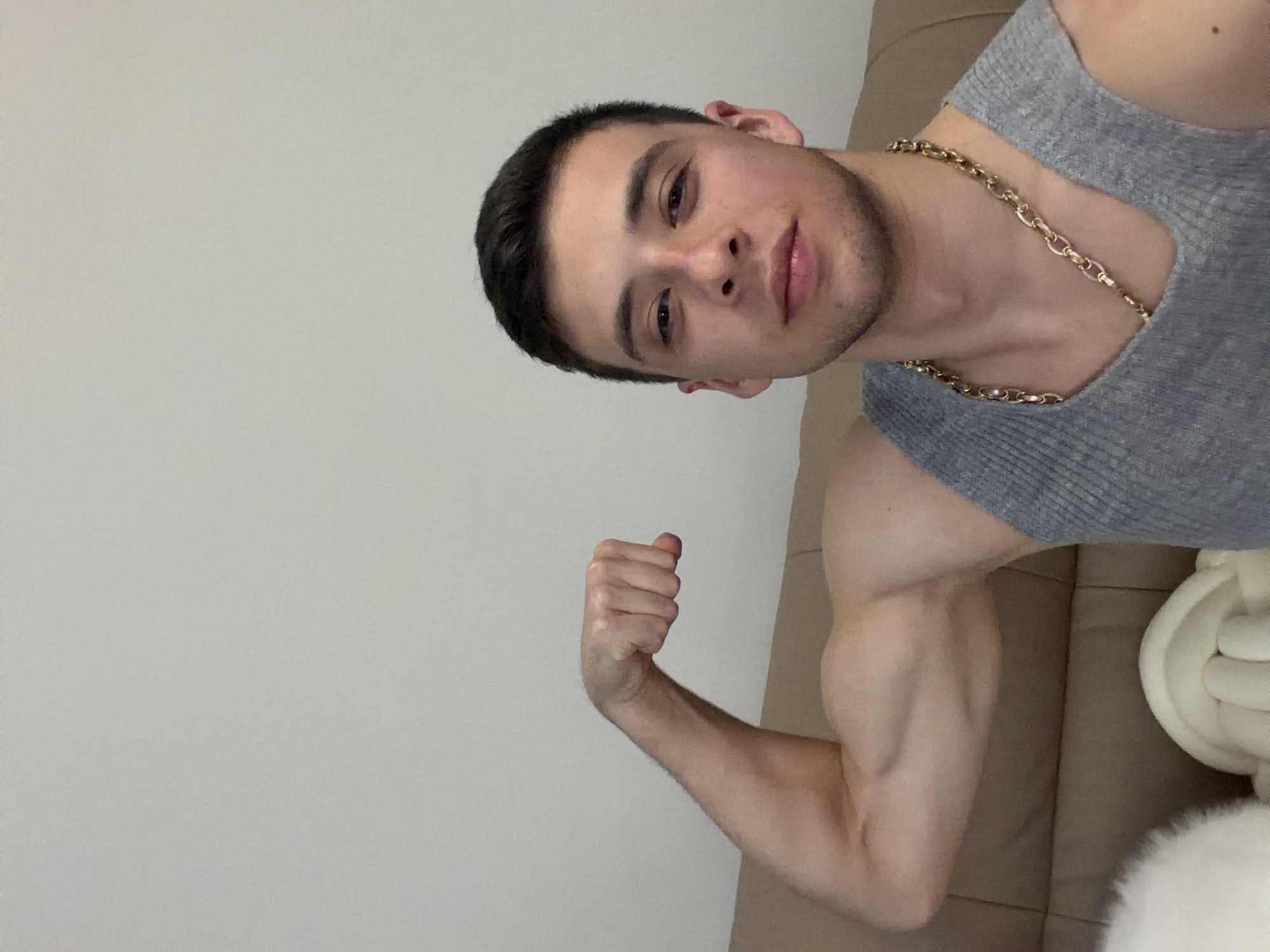 The image shows a young man with short dark hair, wearing a gray tank top and a gold chain, flexing his left bicep. He is posed against a light beige wall and a couch of similar color, with a white decorative knot pillow partially visible. The location is Unknown, Unknown.