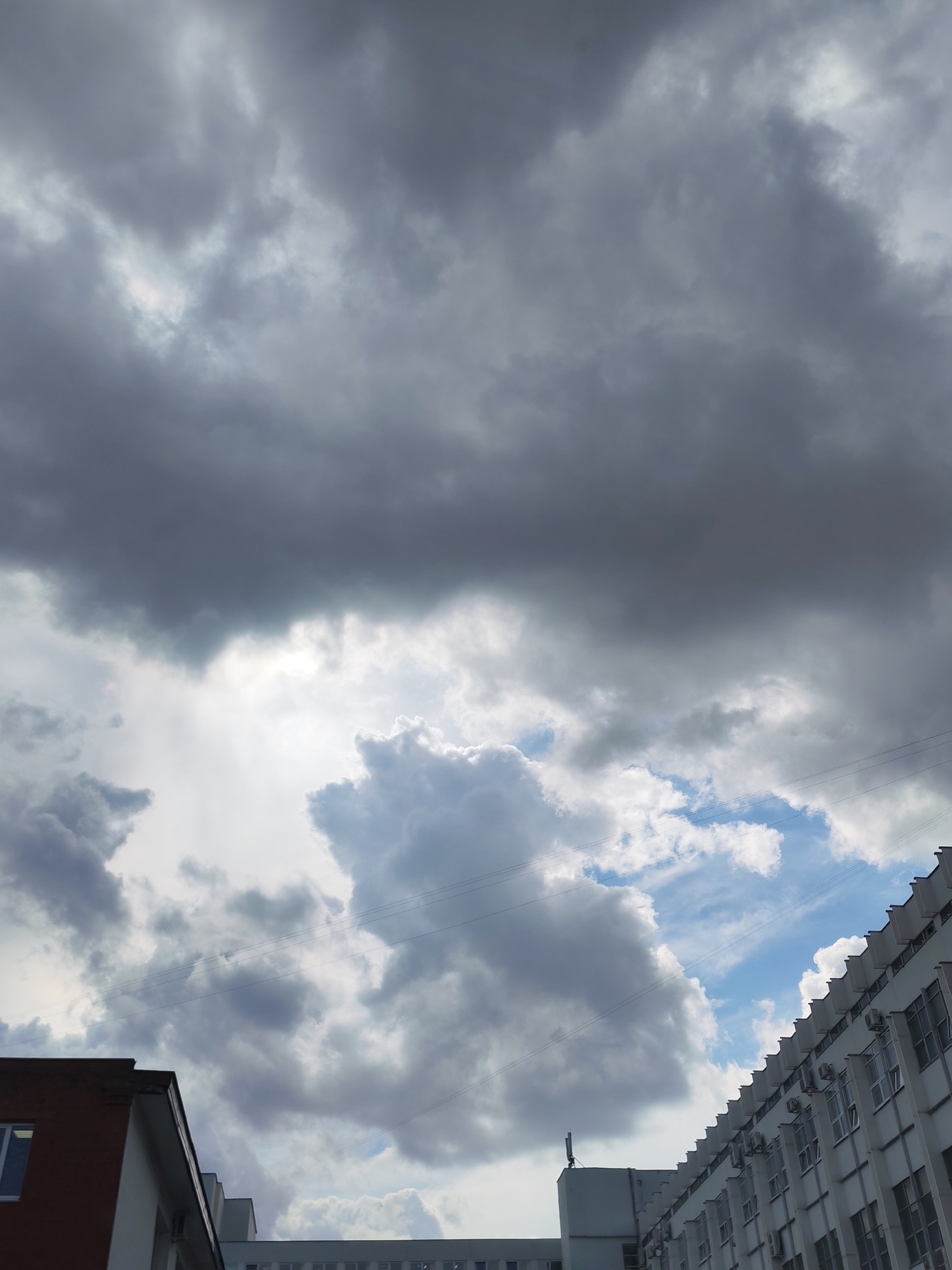 An outdoor, upward-angled shot captures a dramatic sky dominating the frame, with sections of urban buildings at the bottom. The upper portion of the sky is filled with dense, dark grey clouds, suggesting an overcast or stormy condition. Below these, bright light breaks through lighter grey and white clouds, indicating direct sunlight partially obscured. Towards the bottom right, patches of clear blue sky are visible amidst fluffy white clouds. Several thin, dark power lines stretch horizontally across the mid-section of the sky.
On the lower left, a portion of a building with a reddish-brown brick facade and a white upper section is visible, featuring a window. In the center-bottom, another white building with a flat roof can be seen. On the right, a large, multi-story white building extends upwards and to the right, featuring numerous windows and a distinctive roofline adorned with a series of small, repetitive projecting structures, each with an air conditioning unit or similar fixture mounted below it. The architecture suggests a functional or administrative building, possibly an industrial complex. The scene is set during the daytime in Izhevsk, Russia. No people or visible text are present.