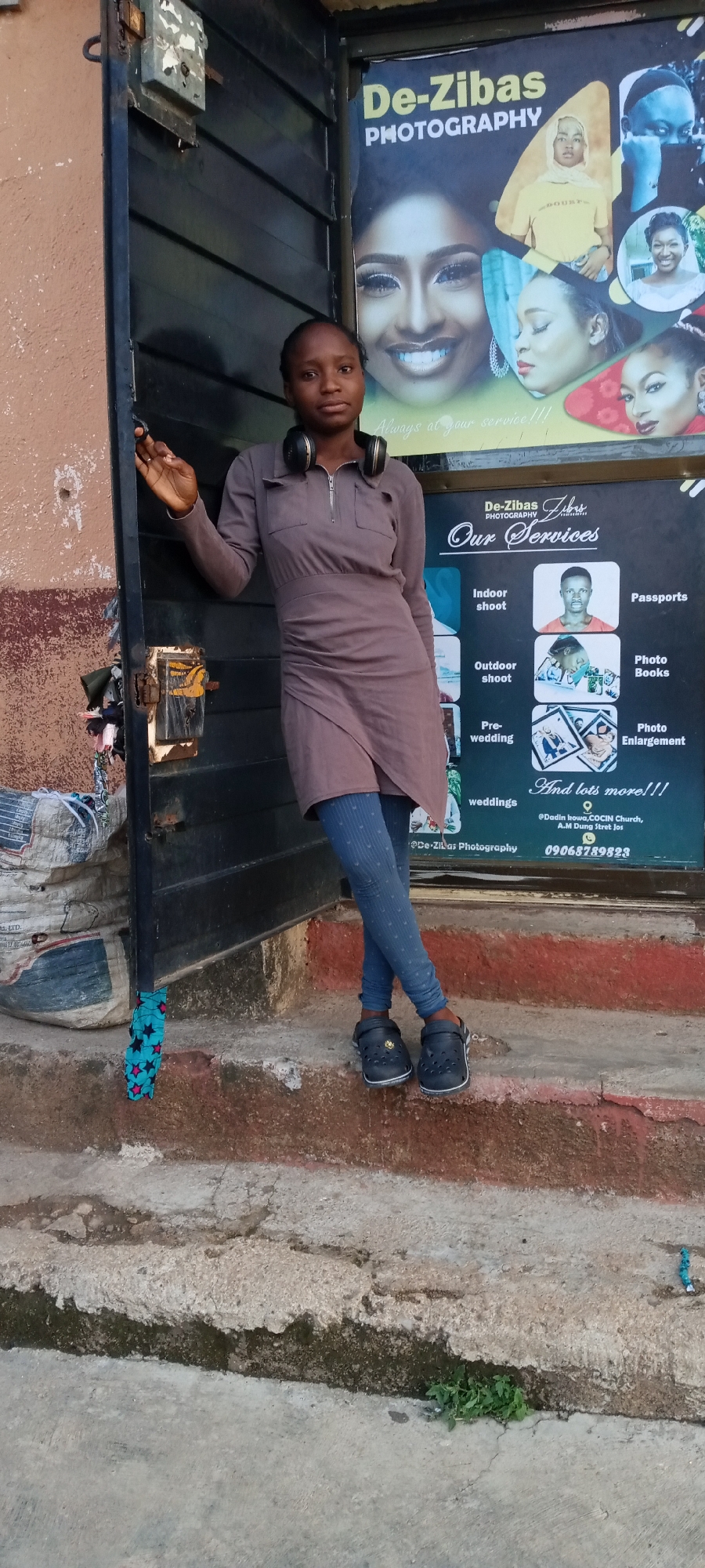 A young woman stands posed in front of a photography studio in Jos, Nigeria. She is positioned on a worn concrete step, leaning against a dark brown, possibly metallic, door frame. She wears a long-sleeved, collared, light brown tunic dress with a zipper at the neckline and pockets, paired with fitted blue leggings featuring a subtle pattern, and dark Crocs shoes. Black over-ear headphones rest around her neck. She has dark hair styled close to her head and looks directly at the viewer with a neutral expression.
To her right, a large banner advertising "De-Zibas PHOTOGRAPHY" is displayed. The banner features portraits of various individuals, including a prominent image of a smiling woman with elaborate makeup. The studio's motto, "Always at your service!!!", is visible. Below this, a section titled "Our Services" lists: "Indoor shoot," "Outdoor shoot," "Pre-wedding," "weddings," "Passports," "Photo Books," and "Photo Enlargement," concluding with "And lots more!!!". The banner also provides a physical address: "@Dadin howa, COCIN Church, A.M Dung Stret Jos," and a phone number: "09068789823."
The immediate surroundings include a rustic, light brown wall to her left, a large bundled sack at the base of the door, and several cracked concrete steps leading down to the ground. Small green weeds are visible growing from cracks in the ground. The lighting suggests it is daytime.