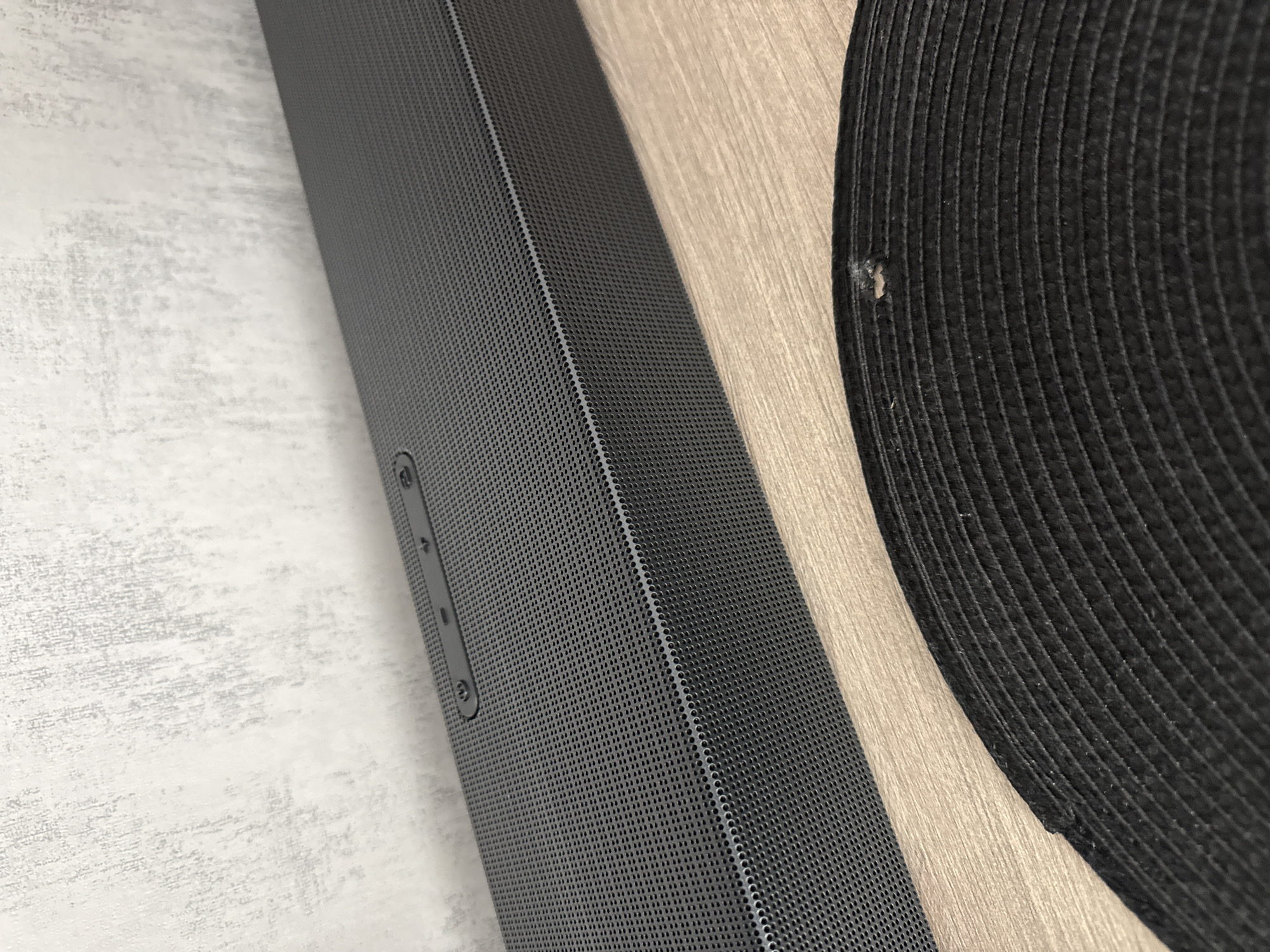The image shows a close-up view of a dark gray soundbar resting on a surface with a light wood-grain texture. A round, dark-colored object with a ribbed or textured surface is partially visible to the right of the soundbar. The surface under the soundbar appears to be a table or countertop. Part of a wall is visible on the left side of the soundbar, featuring a light gray textured pattern. The soundbar has a fine perforated grill and some visible buttons on the left side. The overall lighting suggests an indoor setting. There is no indication of the location being Lovech, Bulgaria, or any people.