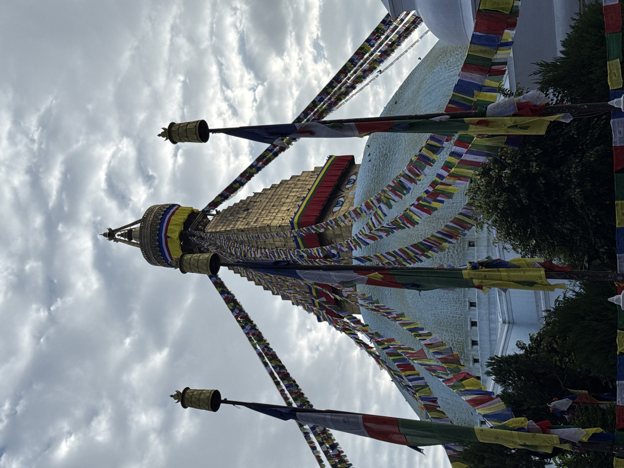 The image depicts a low-angle view of the Boudhanath Stupa, a prominent landmark in Kathmandu, Nepal. The stupa is characterized by its golden pyramidal spire, adorned with prayer flags in various colors that are strung across its surface and extend outwards. Two tall poles with cylindrical decorative elements at their apex are visible in the foreground, also draped with prayer flags and larger prayer banners. The stupa itself has a distinctive face painted on its upper section. The sky above is overcast with scattered clouds, and there are some trees and other structures visible at the base of the stupa. The overall scene captures the spiritual and cultural atmosphere of this important Buddhist site.