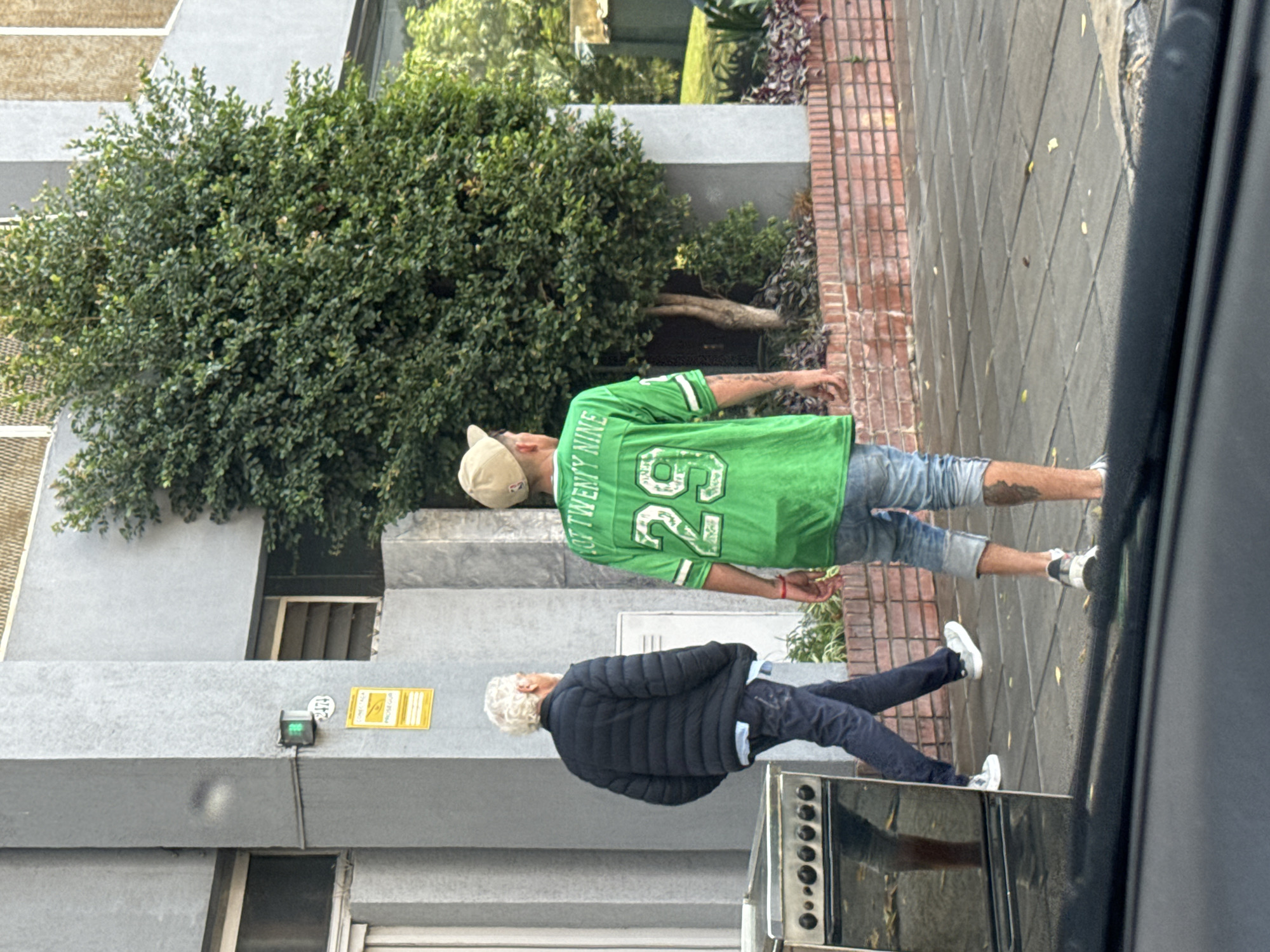 The image shows a street scene. Two men are walking on a sidewalk. One man is wearing a green shirt with the number 26 and the words "TWENTY NINE". He also wears jean shorts, a baseball cap, and white sneakers. The other man is wearing a black jacket and blue jeans, and white sneakers. They are walking next to a building, in front of a large bush. The image was taken from inside a car. The location is likely Buenos Aires, Argentina.