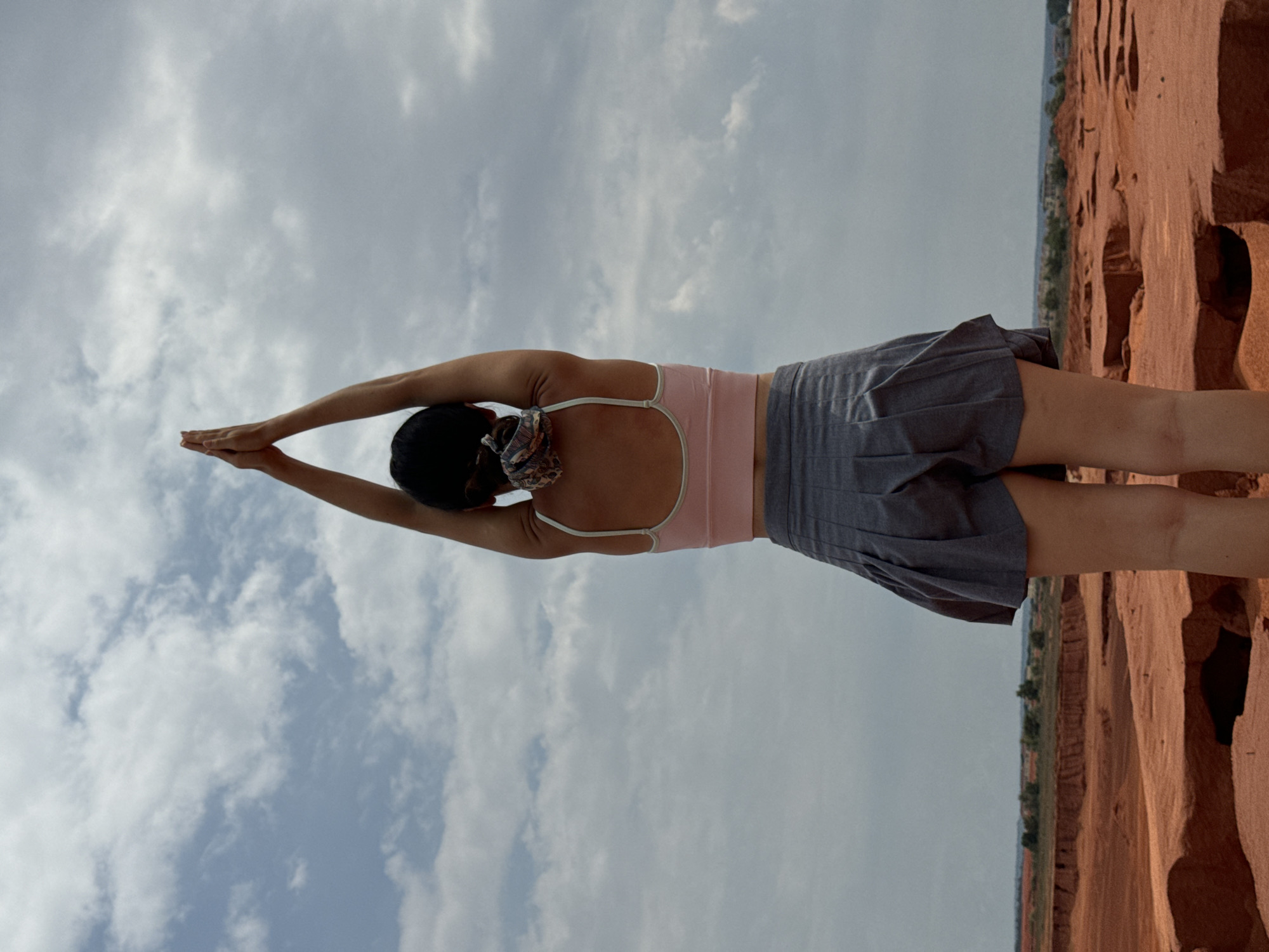 A person stands in a yoga pose with arms extended upward and hands clasped in front of a cloudy sky. The person is wearing a pink sports bra and a gray pleated skirt. The background shows a reddish, arid landscape with some vegetation and buildings in the distance. The location is identified as Mui Ne, Vietnam, a coastal resort town known for its sand dunes.