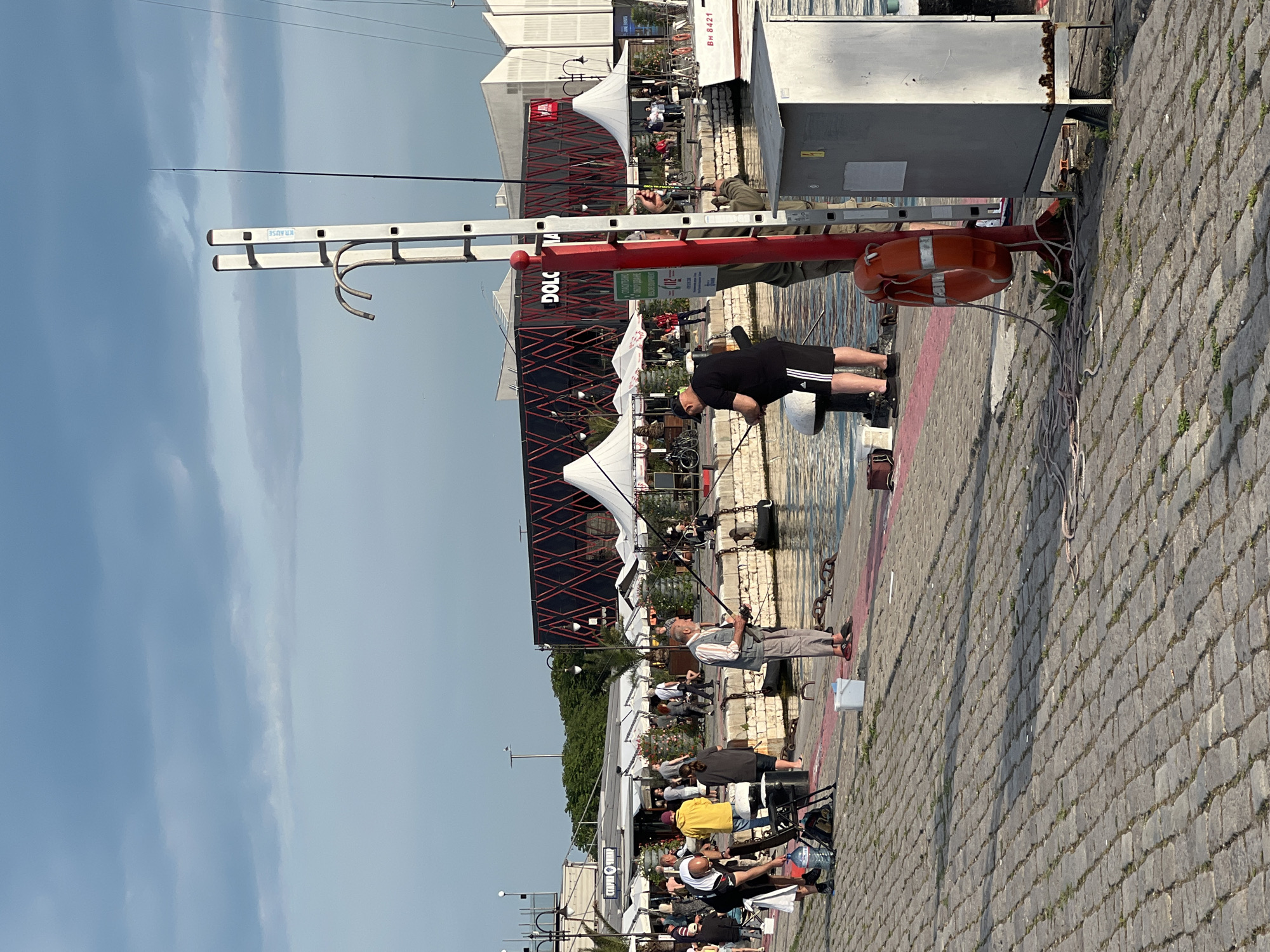 This image captures a vibrant daytime scene along a cobbled waterfront, likely a pier or marina in Varna, Bulgaria. The sky is clear blue with faint wisps of clouds, suggesting pleasant weather.
In the midground, several people are engaged in fishing. One man, wearing a black t-shirt and shorts with a cap, is bending over, attending to his fishing rod. Another man, dressed in a striped shirt and light pants, stands further back, holding a fishing rod over the water. Numerous fishing rods are set up along the edge of the pier.
Prominently featured in the center is a tall, red pole structure with a white top, possibly a crane or support, to which an orange life buoy is attached. A silver ladder, marked with the text "KRAUSE," leans against this structure. Ropes and cables are visible around the red pole and on the ground. The ground is paved with distinct gray cobblestones, with some weeds growing between them.
In the background, a large, dark building with red geometric accents and the partial text "DOLC" (likely "DOLCE") is visible. White tent-like canopies are set up in front of it, beneath which more people are gathered, possibly dining or socializing at tables and chairs. Some bicycles are parked nearby. A white structure or boat with the text "BH 8421" is partially visible on the right. A green sign attached to the red pole displays Cyrillic text, including the number "112" (an emergency number). A white bucket and a large plastic water bottle are also seen near the fishermen. The overall atmosphere appears relaxed and leisurely.