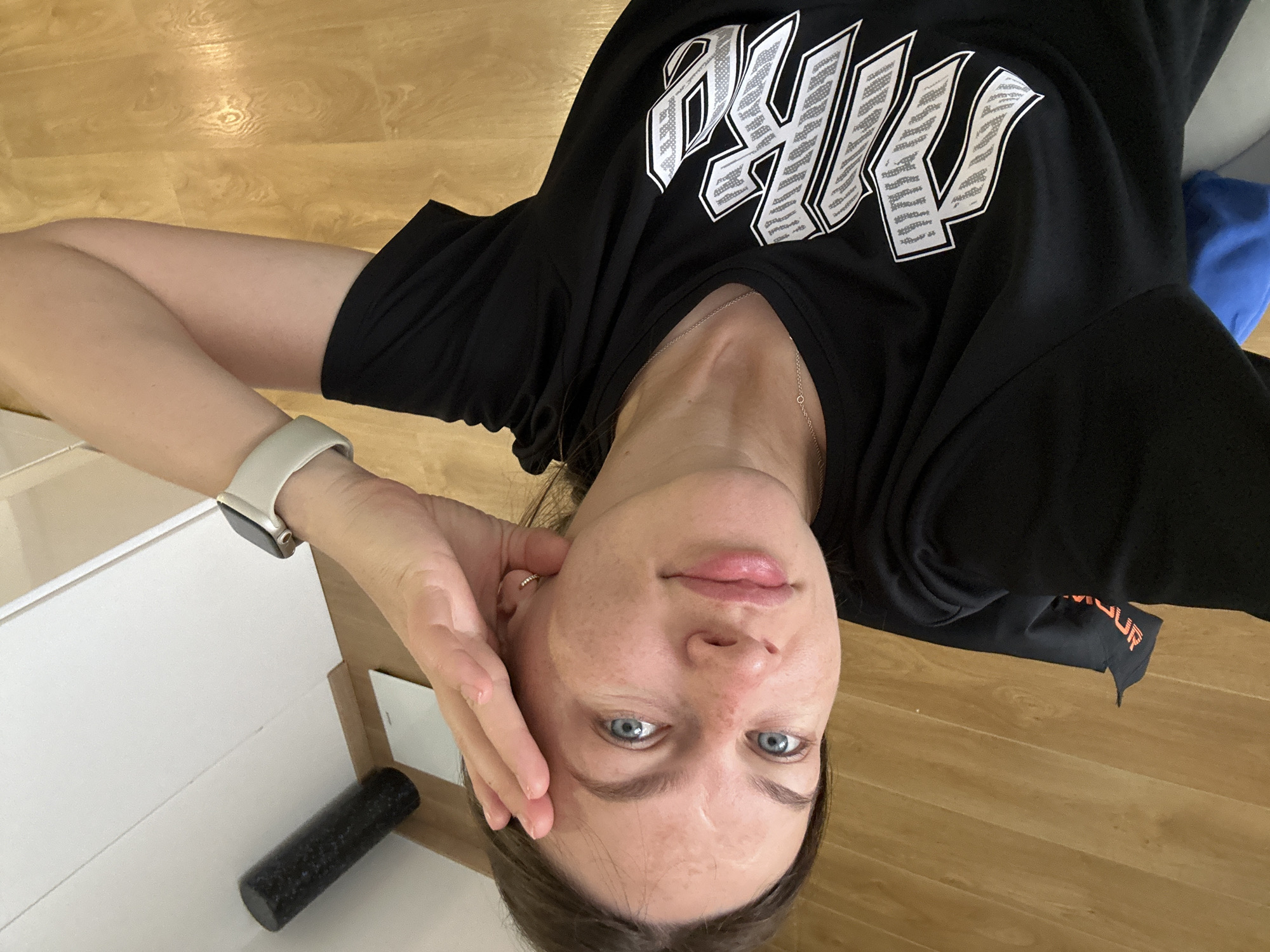A woman with blue eyes is lying on her back on a light brown wooden floor. She is wearing a black t-shirt with white lettering and a silver necklace. Her left hand is near her face, and she has a white smartwatch on her left wrist. Next to her, on a white surface, is a black cylinder. The setting appears to be indoors. There are no indications of the city of Budva, Montenegro in the image.