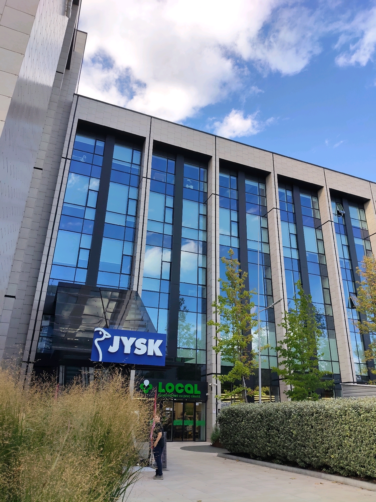 The image shows a modern building with a facade of glass and light-colored stone tiles. The building has multiple floors of large, rectangular windows, reflecting the blue sky and white clouds. In front of the building, there is a blue sign with white lettering that reads "JYSK". To the right of the JYSK sign, a green sign with white lettering reads "LOCAL Supermarket". A person is standing on the sidewalk in front of the supermarket entrance, holding a broom and dustpan, and appears to be cleaning. There are trees with green and yellow leaves planted along the sidewalk, and a well-maintained hedge runs along the edge of the building. The scene appears to be a commercial area within the city of Chișinău, Moldova, likely a shopping center or retail park.