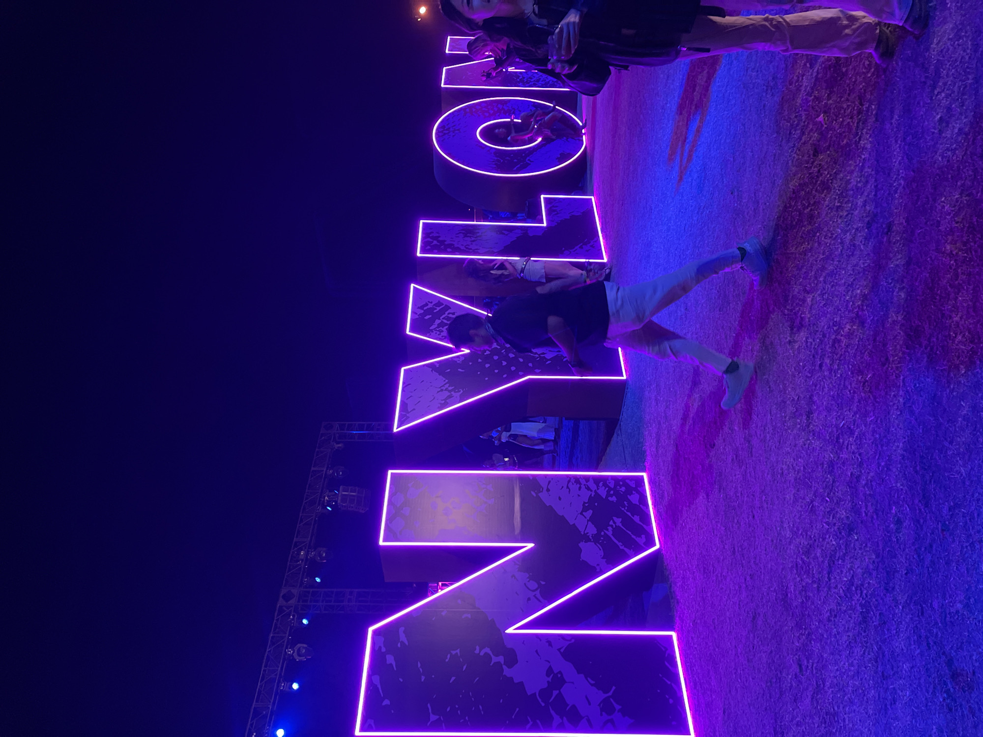 The image shows a large neon sign spelling "NYLON," likely at an event. The sign is illuminated with a purple-pink glow. People are standing and posing in front of the sign on what appears to be a grassy lawn. The background is dark, suggesting it's nighttime, with a lighting rig visible on the left side. The location is Thermal, United States of America.