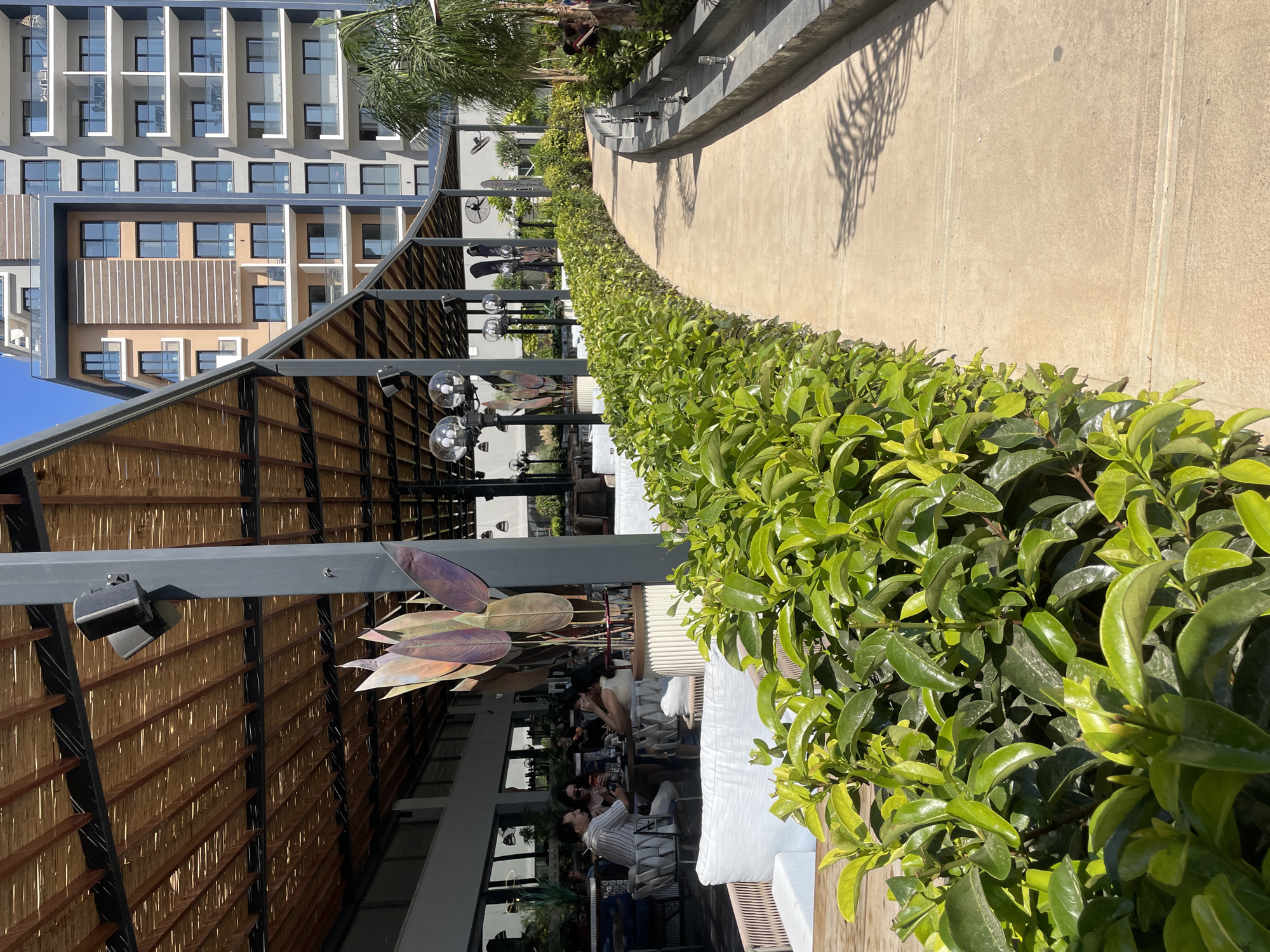 This is a photograph of a contemporary outdoor dining area at a beachfront resort in Trikomo, Northern Cyprus. In the foreground, a curved pathway made of concrete is flanked on the left by lush green bushes and on the right by a low concrete structure with water spouts, suggesting a water feature. A large, modern building with multiple balconies and windows dominates the background. The dining area itself features a slatted bamboo roof supported by dark metal pillars, with patrons seated at tables beneath. The overall scene conveys a sense of relaxed luxury and a vacation setting.