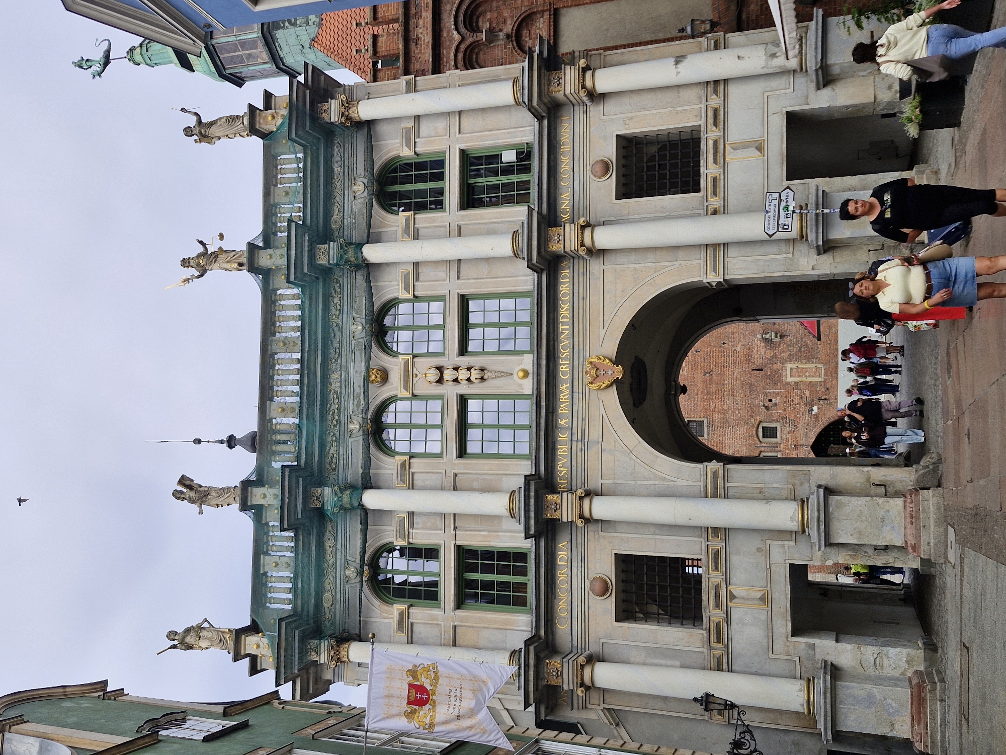 This is a full, eye-level, slightly low-angle, vertical shot of the Golden Gate in Gdańsk, Poland, on a cloudy day. The gate is an elaborate Renaissance-style structure with two main levels. The upper level features a balustrade adorned with statues, and the lower level has a large arched passageway. The gate is decorated with intricate stonework, columns, and gold accents. Above the archway, a coat of arms is visible.
On the left side of the gate, a banner hangs, displaying a crest with lions and a cross. The street in front of the gate is paved with cobblestones. Several people are walking through the archway and in the foreground. In the foreground, from left to right, a woman in a light yellow polo shirt and a denim skirt walks past, followed by a woman in a black t-shirt and black leggings, and then a woman in a beige hooded sweatshirt and blue jeans walks away from the camera. More people are visible walking through the archway in the background, heading towards a brick building. A street sign pointing to "GDANSKI TEATR SZKECIOWY" (Gdańsk Puppet Theatre) and "CENTRUM" (Center) is mounted on a pole to the right of the archway. The sky is overcast and a single bird is visible flying in the upper center of the frame.