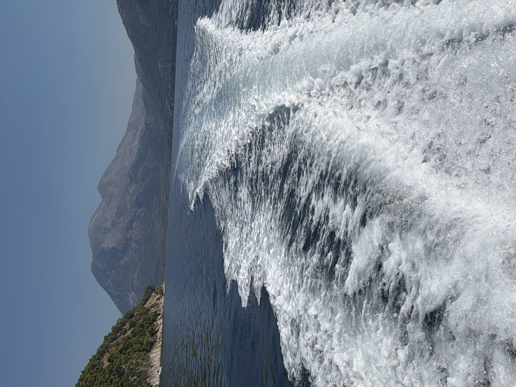 The image shows a wake trailing behind a boat moving through dark blue water. The wake is a frothy white, with smaller bubbles and sparkling light reflecting off the surface. In the distance, the blue sea stretches towards a coastline with rolling hills covered in dense green vegetation. Further inland, larger, rugged mountains rise against a clear blue sky. The sun is bright, casting a sharp light on the scene. This vista is consistent with the coastal areas of the Municipal Unit of Kalamos, Greece, which features a mountainous interior and a shoreline on the Ionian Sea.