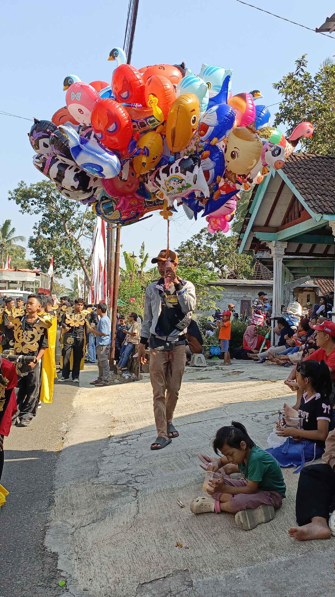 The image depicts an outdoor scene in Krajan Lor, Indonesia, on a sunny day, likely during a local festival or event. A man in a brown cap, grey jacket, and brown pants is walking, holding a large cluster of colorful balloons aloft. The balloons are shaped like various cartoon characters, animals, and objects. In the foreground, to the right, a young girl in a green shirt and purple pants sits on the ground, holding a clear plastic cup and appearing to eat or drink. Other people, including adults and children, are visible in the background, some seated and others standing, observing the event. A group of young men in traditional attire, adorned with gold and black costumes and yellow sashes, are marching or parading on the left side of the image. The environment includes a paved road, concrete structures, trees, and a building with a traditional Indonesian architectural style. Red and white flags are also visible, suggesting a national celebration or commemoration.