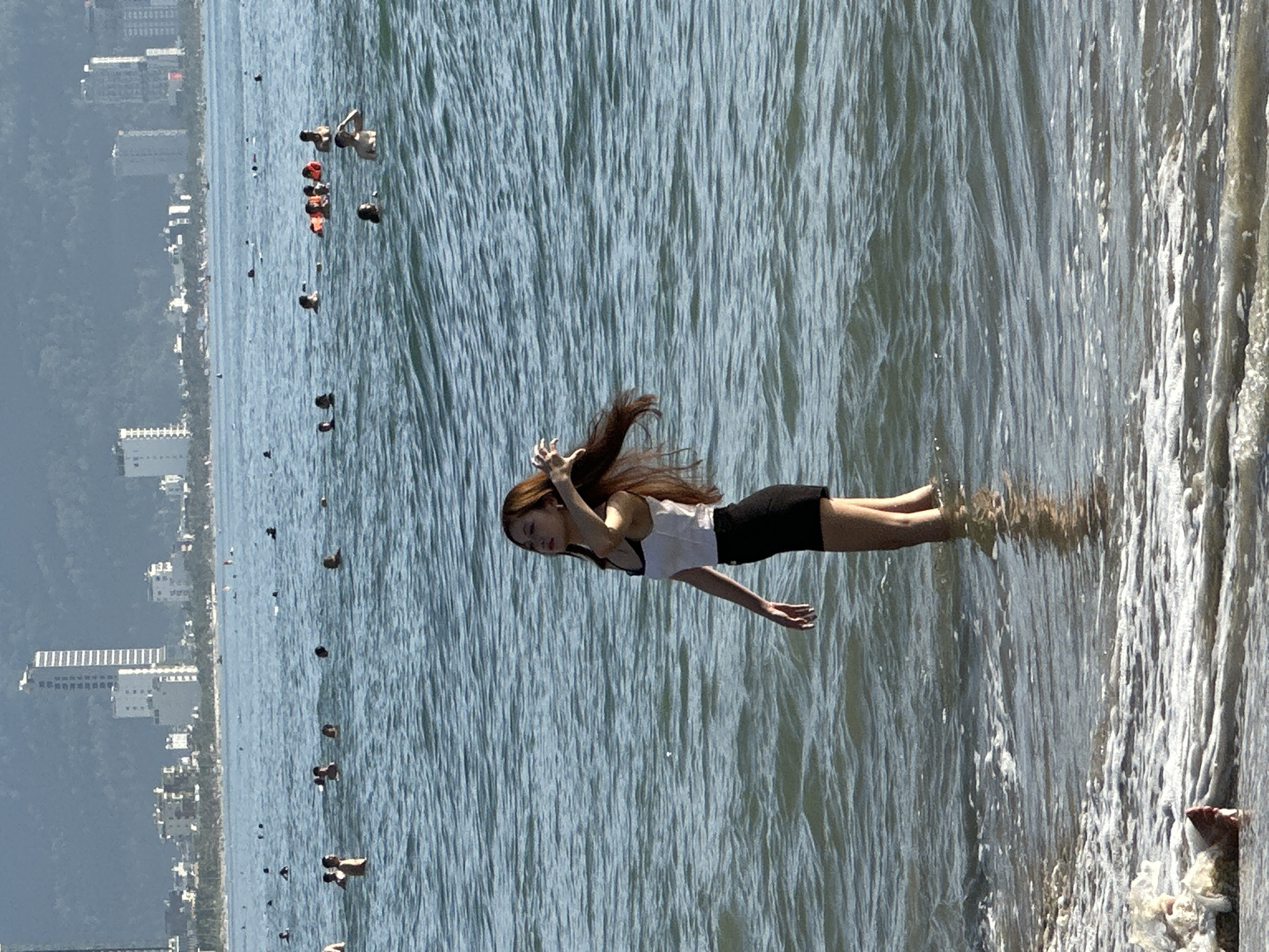 An adult female with long, dark hair is standing in shallow water at My Khe Beach in Da Nang, Vietnam. She is wearing a white tank top and a black skirt. The ocean is calm with gentle waves. In the background, a city skyline with tall buildings is visible. Several other people are in the water, swimming and enjoying the beach. The sun is shining, casting a warm light on the scene.