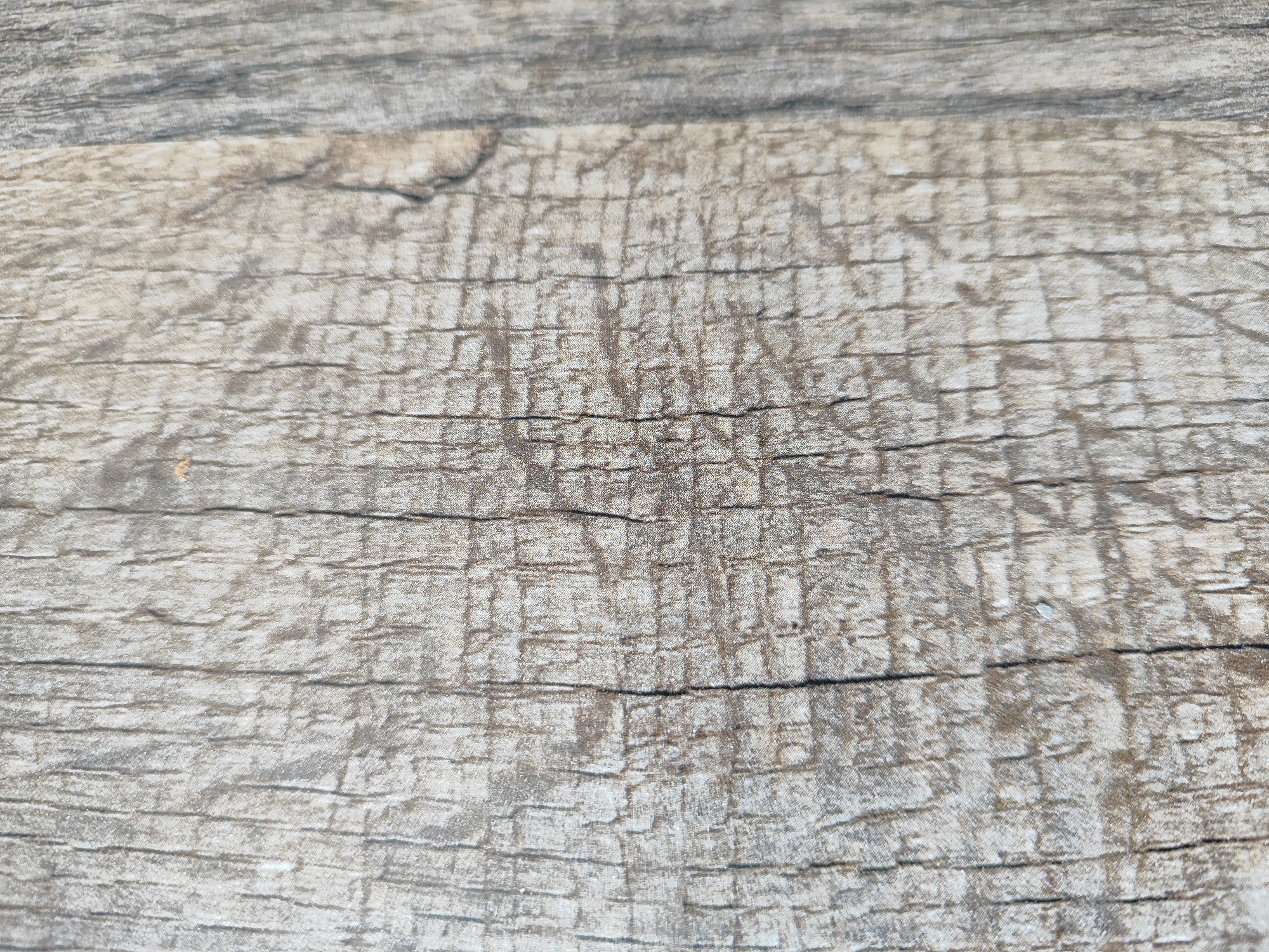 This is a close-up, overhead shot of a weathered wooden surface. The wood grain is prominent, with horizontal lines and darker cracks running throughout. The color palette consists of muted grays and browns, suggesting natural aging or a specific finish. The texture appears rough and possibly textured, with some areas looking lighter and more reflective, while others are darker and more matte. There are no people, objects, or identifiable scene elements present. The image focuses solely on the texture and pattern of the wood. No text is visible. The lighting appears to be natural, possibly from an overcast sky, as there are no strong shadows. The overall impression is one of natural, aged material.