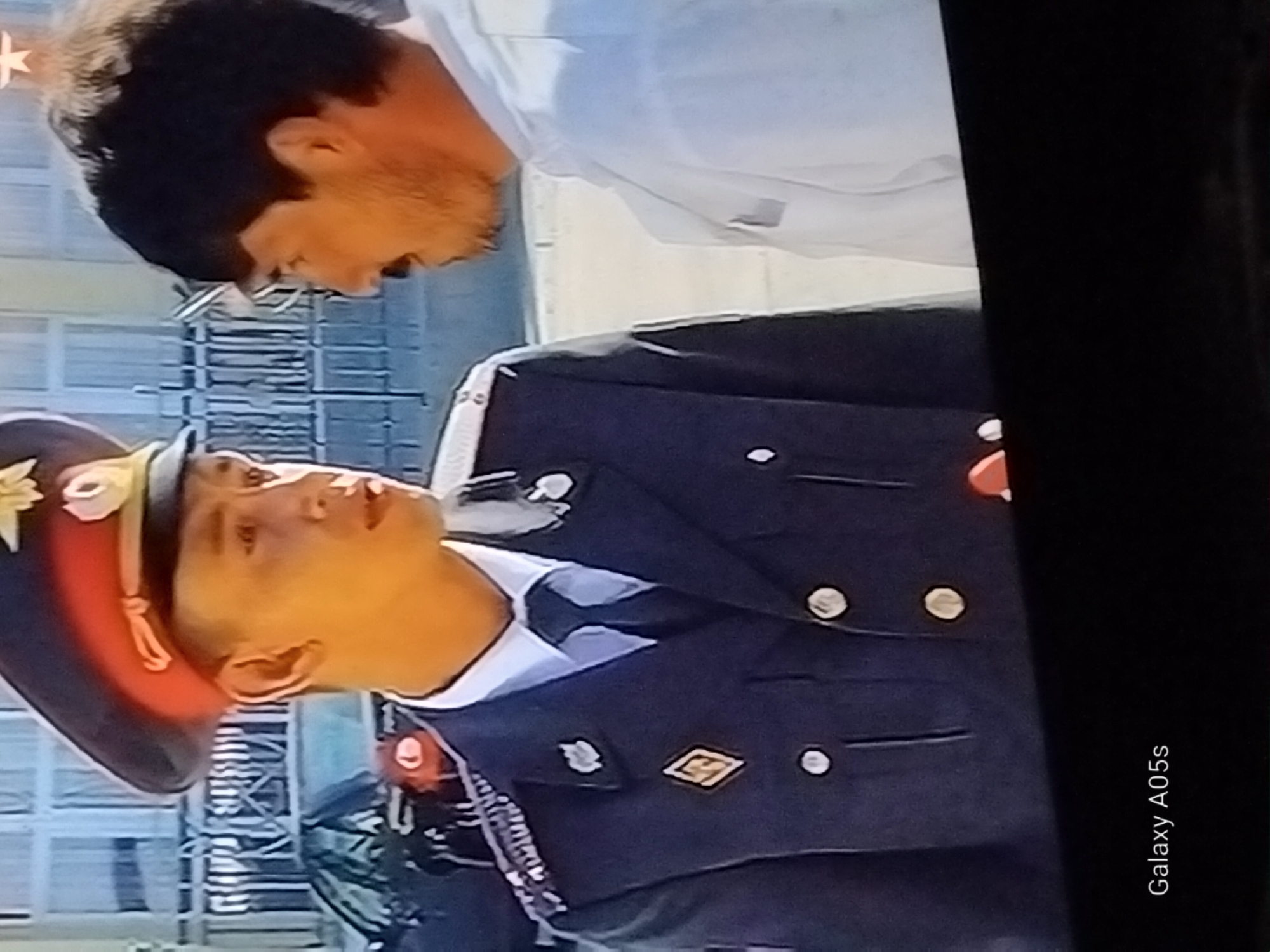 The image shows two men in an outdoor urban setting, possibly the city of Druzhba, Russia. On the left, a man in a dark police uniform with a red and gold hat is speaking to another man. The police uniform features insignia on the collar and shoulder, and a diamond-shaped badge on the chest. The man on the right is wearing a white shirt. In the background, a black car and a metal fence are visible. The overall scene appears to be a moment captured from a film or television program.