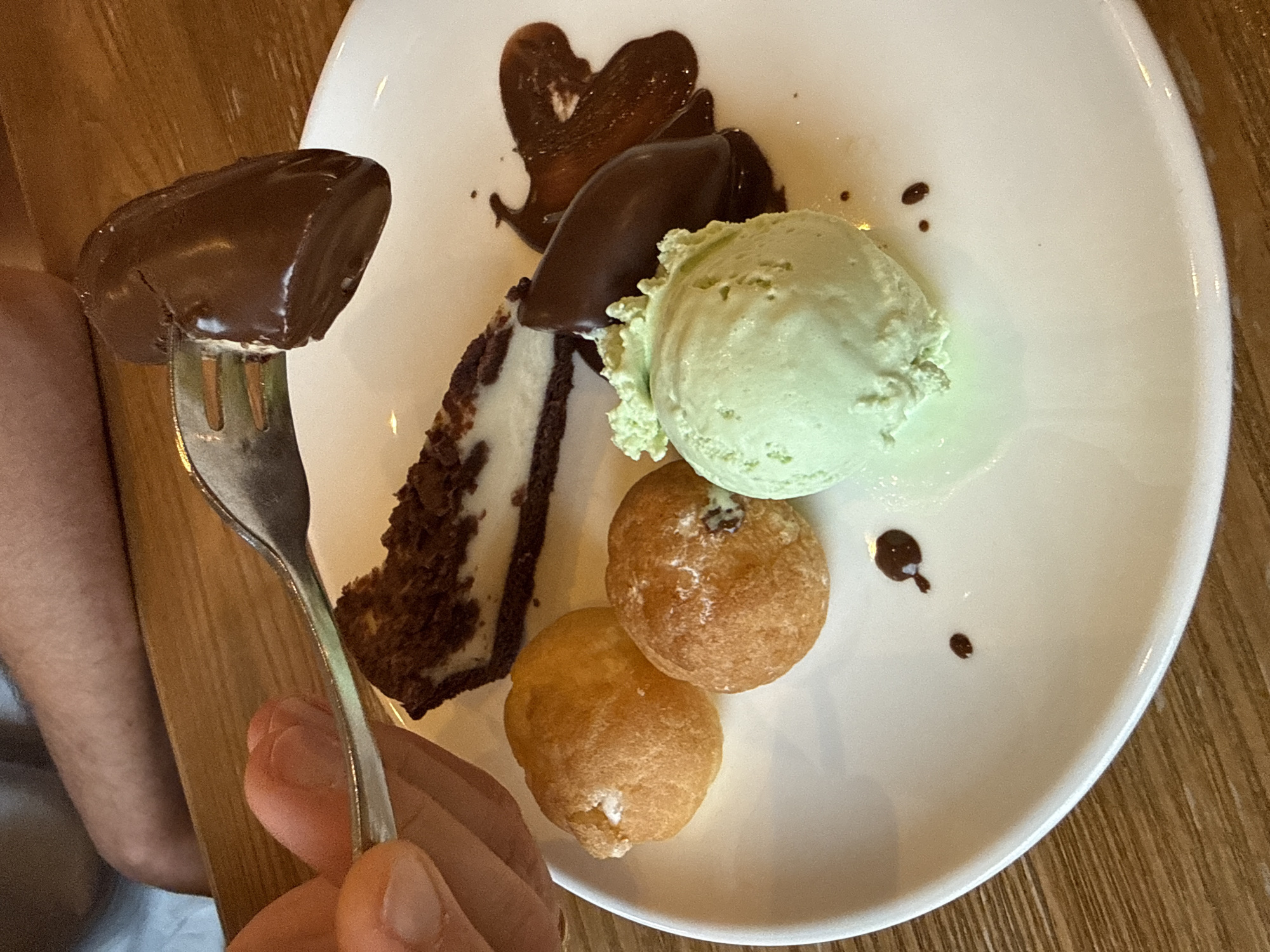 In Nordhausen, Germany, on a white circular plate placed on a wooden surface, a dessert assortment is presented. The plate holds a slice of chocolate cheesecake, a scoop of mint green ice cream, two small, round cream puffs, and a chocolate-covered pastry. A fork, held by a person's hand, is lifting a piece of the chocolate-covered pastry, with a sliver of white visible underneath the chocolate. Drizzled chocolate sauce is spread across the plate, and a small, dark chocolate-colored object, possibly a candy, sits near the ice cream.