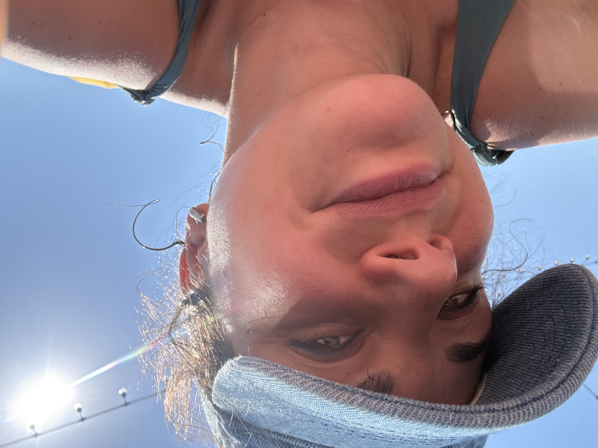 A close-up, low-angle shot depicts a person with their face and upper body angled toward the camera. They are wearing a blue denim hat and what appears to be a teal swimsuit top. Part of a string of lights is visible in the background, illuminated by sunlight. The background is light blue, suggesting the sky. The photograph does not contain any information that would confirm a location in Moscow, Russia.