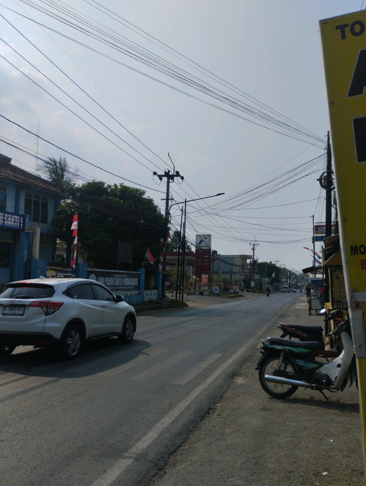 The image depicts a street scene in Purwakarta, Indonesia. A white Honda HR-V is driving on the left side of the road. On the left side of the road, there is a blue building with the text "YAYASAN ISLAM SANTUNG" and "SMK PRABU SAKTI 1 PURWAKARTA" visible. Indonesian flags are displayed on poles in front of the building. On the right side of the road, there are several motorcycles parked on the sidewalk, and a yellow sign with black lettering is partially visible. A Pertamina gas station is visible in the distance on the left side of the road. The sky is overcast, and numerous power lines crisscross the scene.