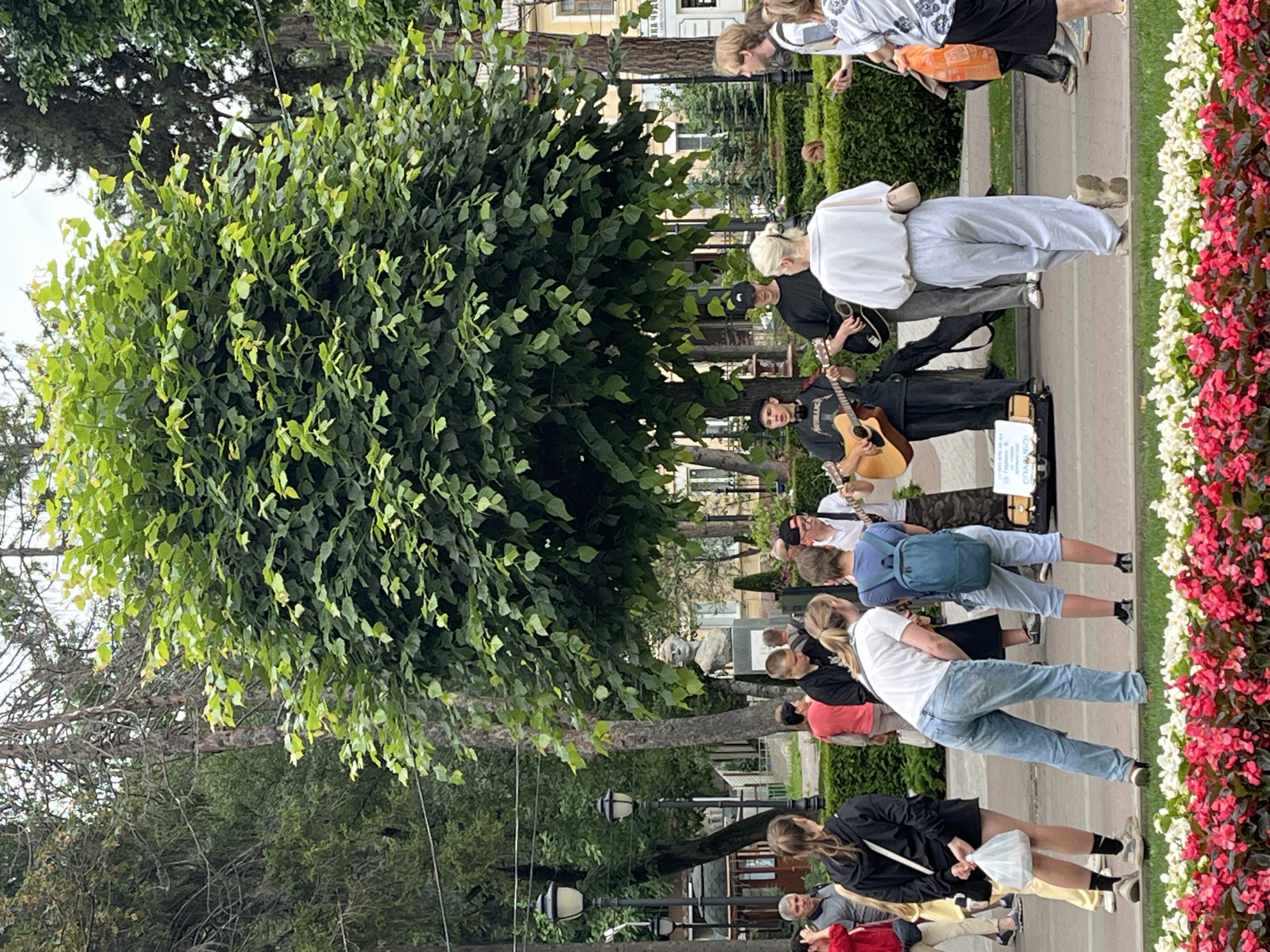 The image captures a street scene in Kislovodsk, Russia. In the foreground, a vibrant bed of red and white flowers lines a paved walkway. Beyond the flowers, several people are gathered, observing a group of street musicians. Two young men are playing acoustic guitars and singing, with a third individual standing nearby. The musicians are set up with an open guitar case on the ground, presumably for donations. Behind them, a bust of a man stands on a pedestal. The background features trees, lampposts, and buildings, suggesting an urban park or plaza setting. The overall atmosphere appears to be casual and public, with people enjoying the impromptu performance.