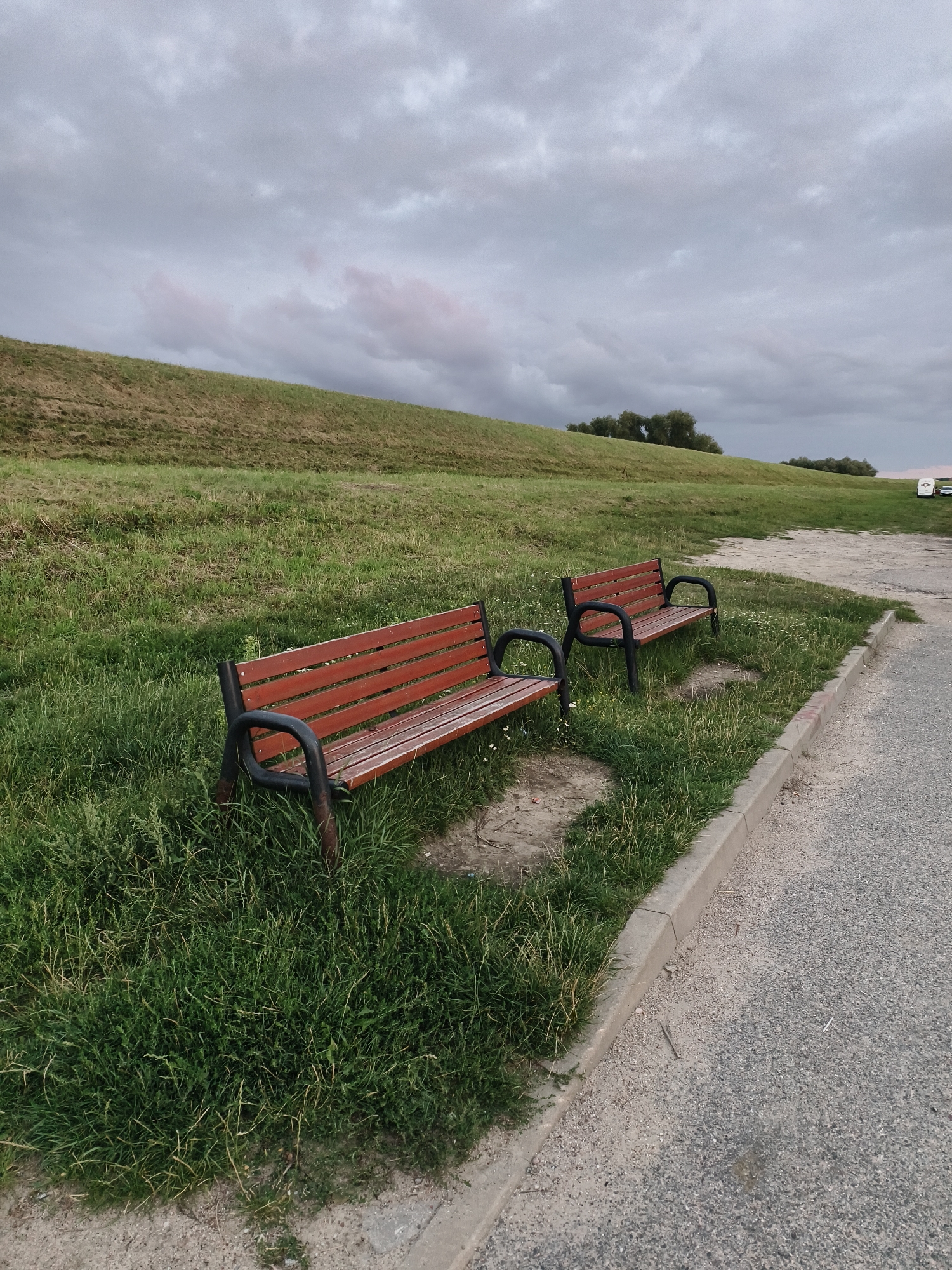 The image depicts an outdoor scene with a sloped grassy hill under a cloudy sky. Two wooden park benches with dark metal frames are situated on the grass near a paved area. The bench in the foreground is closer and angled towards the viewer, while the bench in the background is further away. The grassy slope extends upwards and to the left, with some trees visible in the distance on the right. The paved area consists of gravel and concrete edging. The overall atmosphere suggests a somewhat overcast or late afternoon setting. There are no people or animals visible in the image. The location in the city of Tczew, Poland, is not discernible from the visual information alone.