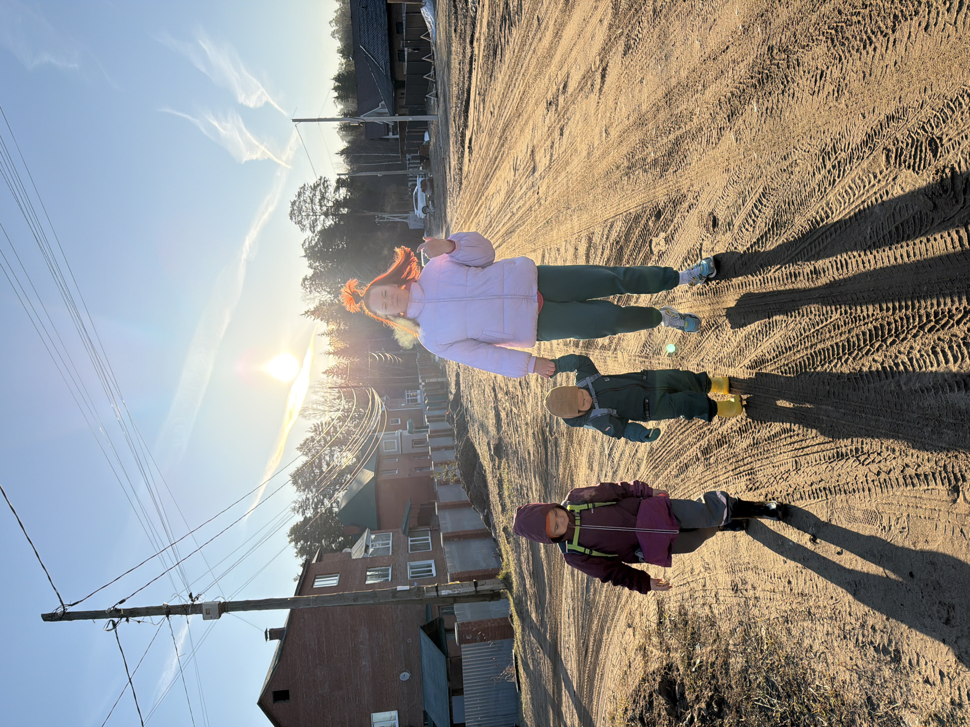 The image shows a dirt road in Chaikovsky, Russia, with tire tracks. A woman wearing a light pink puffer jacket and green pants stands on the road, with two children standing close to her. The child on the bottom wears a purple jacket with a hood and green straps. The other child has on a green jacket, brown hat and yellow rubber boots. The sun is shining brightly, casting long shadows of the people. In the background, there are houses, trees, and power lines.