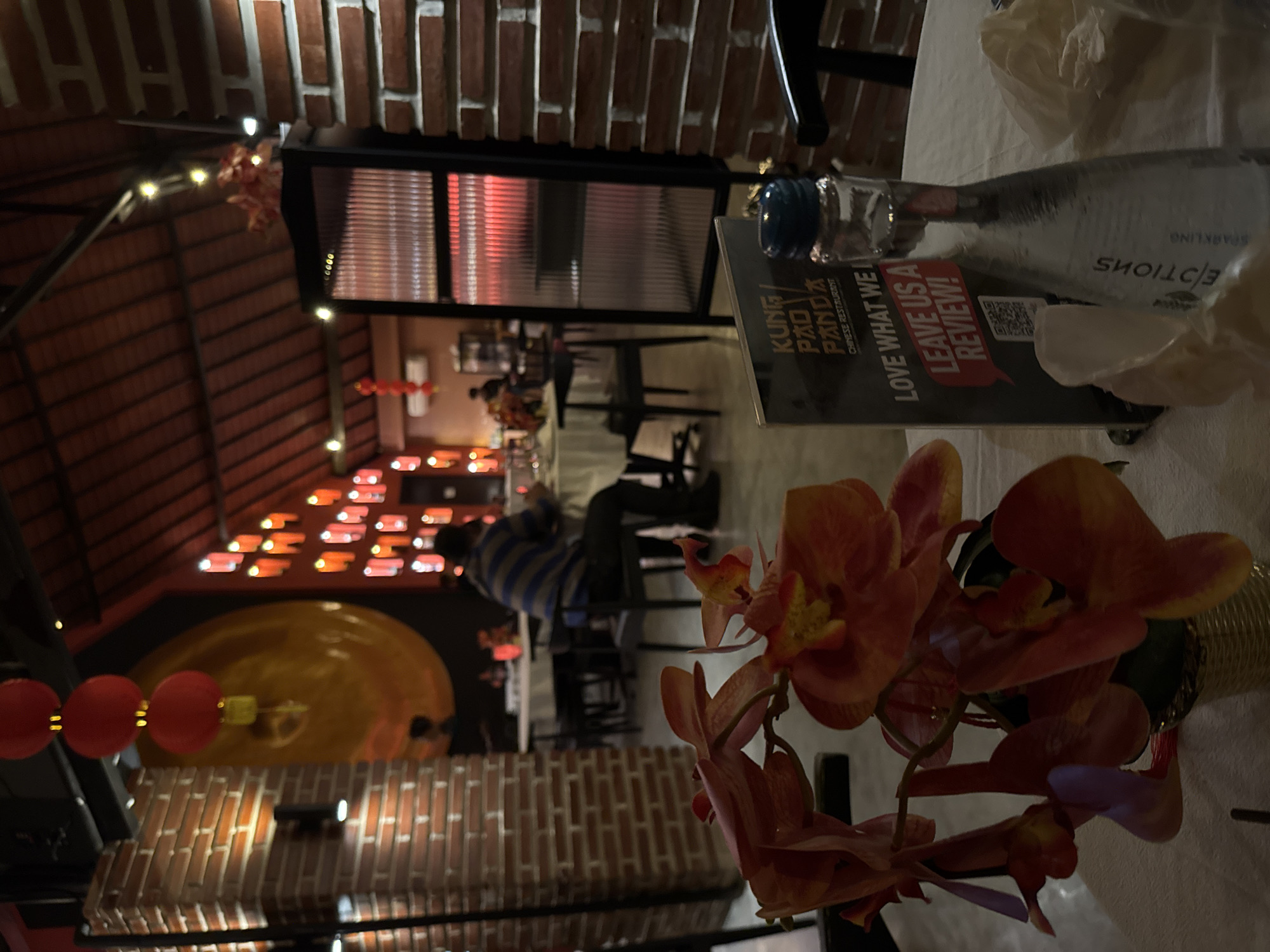 The image depicts the interior of a restaurant, possibly located in Kuta, Indonesia. In the foreground, a close-up view of orange orchids is visible, with part of a table and a bottle of sparkling water. Behind the flowers, a menu card for "Kung Pao Panda Chinese Restaurant" is placed. The restaurant's ambiance is characterized by red lanterns hanging from the ceiling and red brick walls, suggesting an East Asian theme. Several tables with white tablecloths and chairs are arranged throughout the dining area, with patrons seated at some of them. The ceiling features exposed wooden beams and lighting, contributing to a warm and inviting atmosphere.