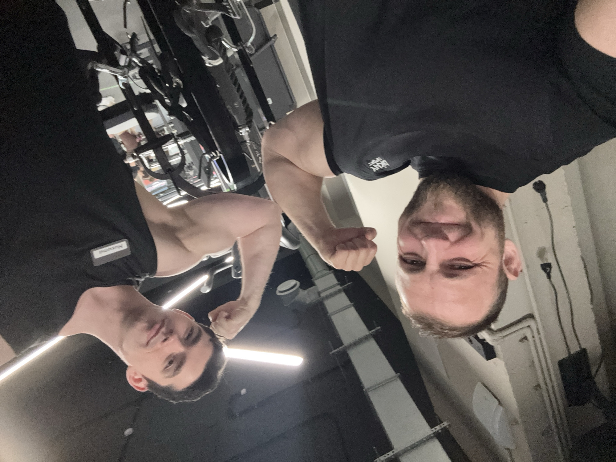 The media file is an upside-down, low-angle selfie featuring two men flexing their biceps in a gym setting.
The man on the right, with a beard, smiles broadly at the camera. He wears a black t-shirt with a "DKNY SPORT" logo visible on his left chest. His right arm is bent, showcasing a flexed bicep.
The man on the left, with dark hair, has a neutral to slightly smirking expression while looking at the camera. He also wears a black t-shirt, which has a white name tag on his right chest, displaying the name "НИКИТА" (Nikita). His left arm is flexed.
The background shows various pieces of gym equipment with metal frames and cables. The ceiling features long, white fluorescent light fixtures and exposed industrial-style pipes or ducts, confirming an indoor fitness facility. The image captures a lighthearted moment, with both subjects posing playfully. The photograph appears to have been taken from a very low vantage point, possibly from the floor, looking up at the men.