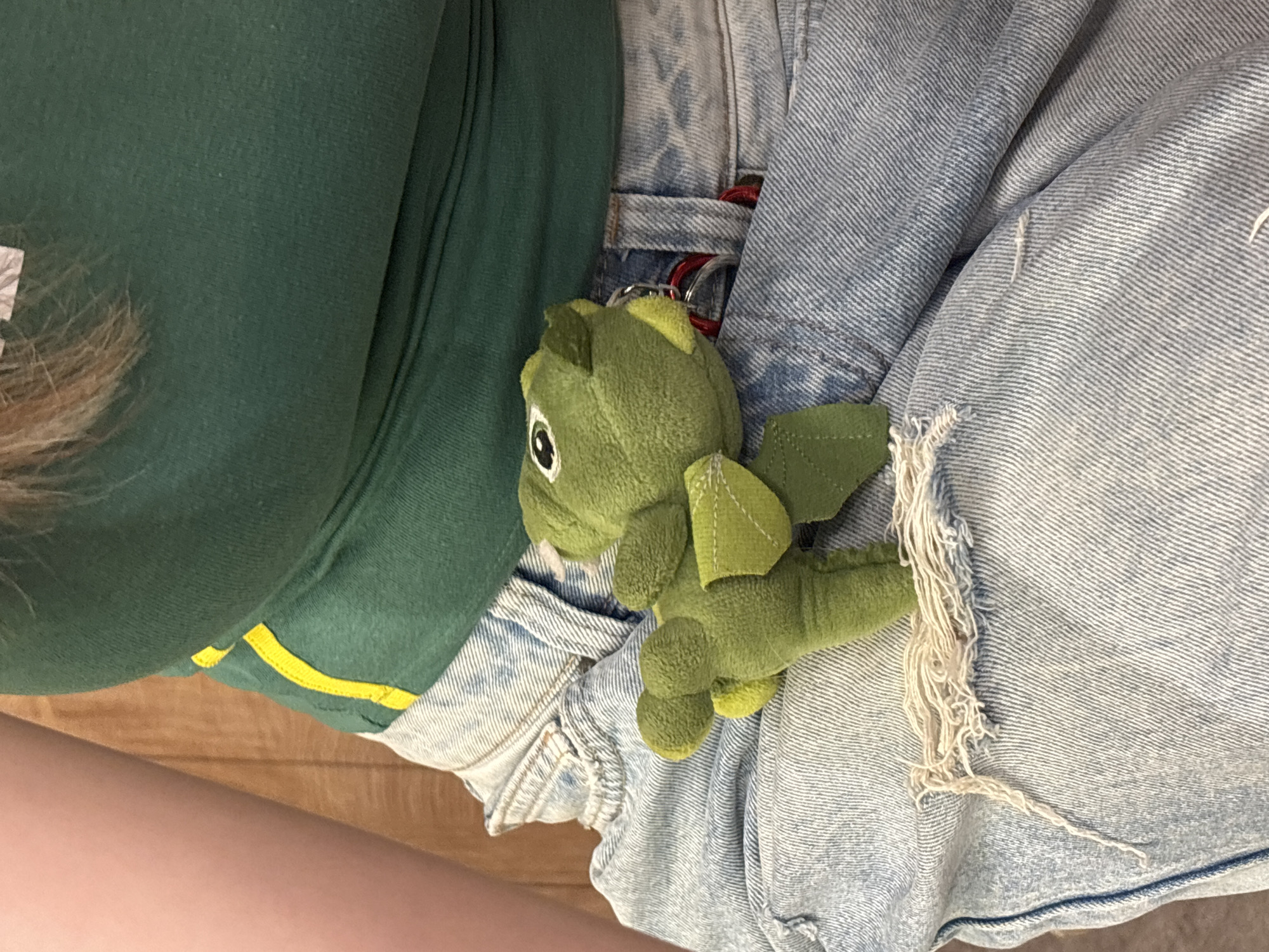 A close-up, low-angle, full-body shot shows a person in light-wash denim jeans with visible rips and fraying, and a green tank top with a yellow stripe. A small, green plush toy dinosaur with wings and light green accents is attached to the jeans via a red lanyard and a silver keyring. The background consists of a wooden surface and a sliver of skin from the person's arm. The scene appears to be indoors, possibly a room. No specific location in Podgorica, Montenegro, can be identified from the image.