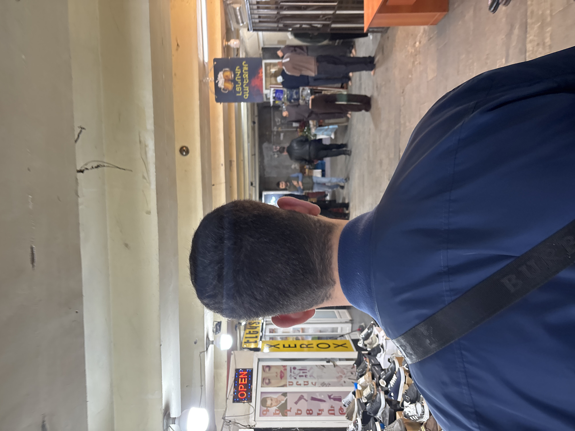 Here is a description of the image:
The image is taken from behind a person wearing a navy-blue jacket and a Burberry strap. The person is in what seems to be a shop or market area. In the background, there are people walking in an alleyway with shops and signs in Armenian. A sign with Armenian script hangs above the scene, possibly an advertisement for "Tigran Mets" beer and snacks.
In the immediate foreground, shelves of shoes and clothing are visible. An "OPEN" sign is lit up, indicating the business is active. Other businesses are visible with signs in Armenian, possibly indicating the location of this image to be in Yerevan, Armenia.
