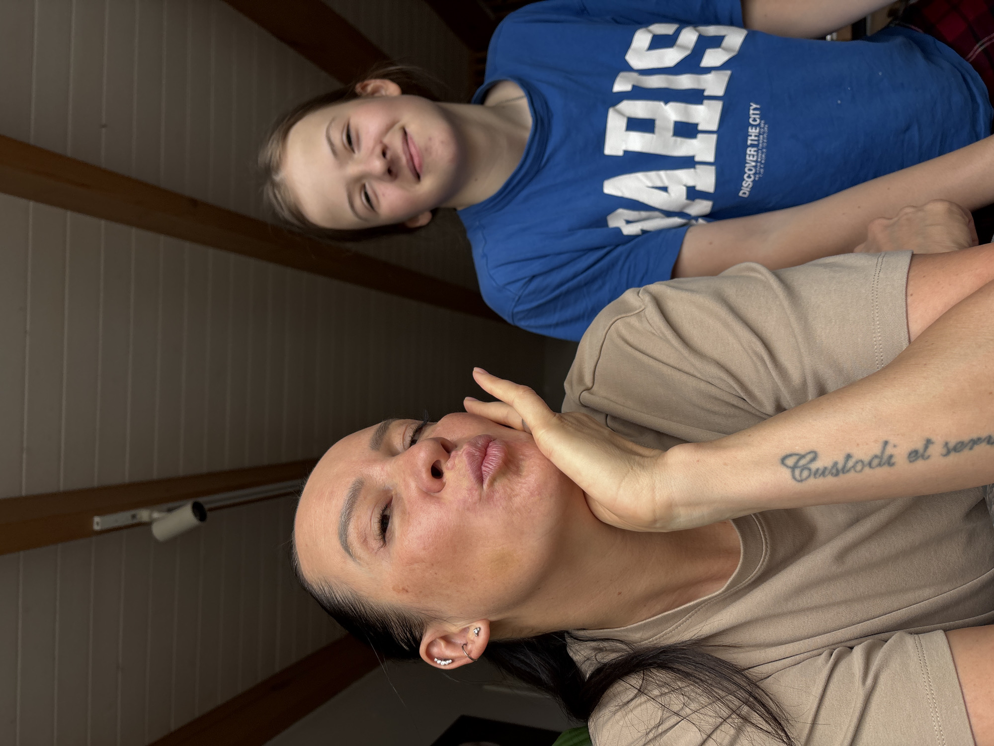 Two individuals are captured in an interior space with a sloped, wood-paneled ceiling. The foreground features an adult female, with dark hair, fair skin and a tattoo on her left forearm, wearing a light brown t-shirt. She is making a kissing face while holding her hand up to her face. Behind her and slightly above is a younger female, wearing a blue t-shirt with white lettering that reads "Paris" and "Discover the City". The location appears to be inside an attic space, indicated by the angled ceiling and wooden beams. It is impossible to provide the exact location in Chaikovsky, Russia from the provided image.