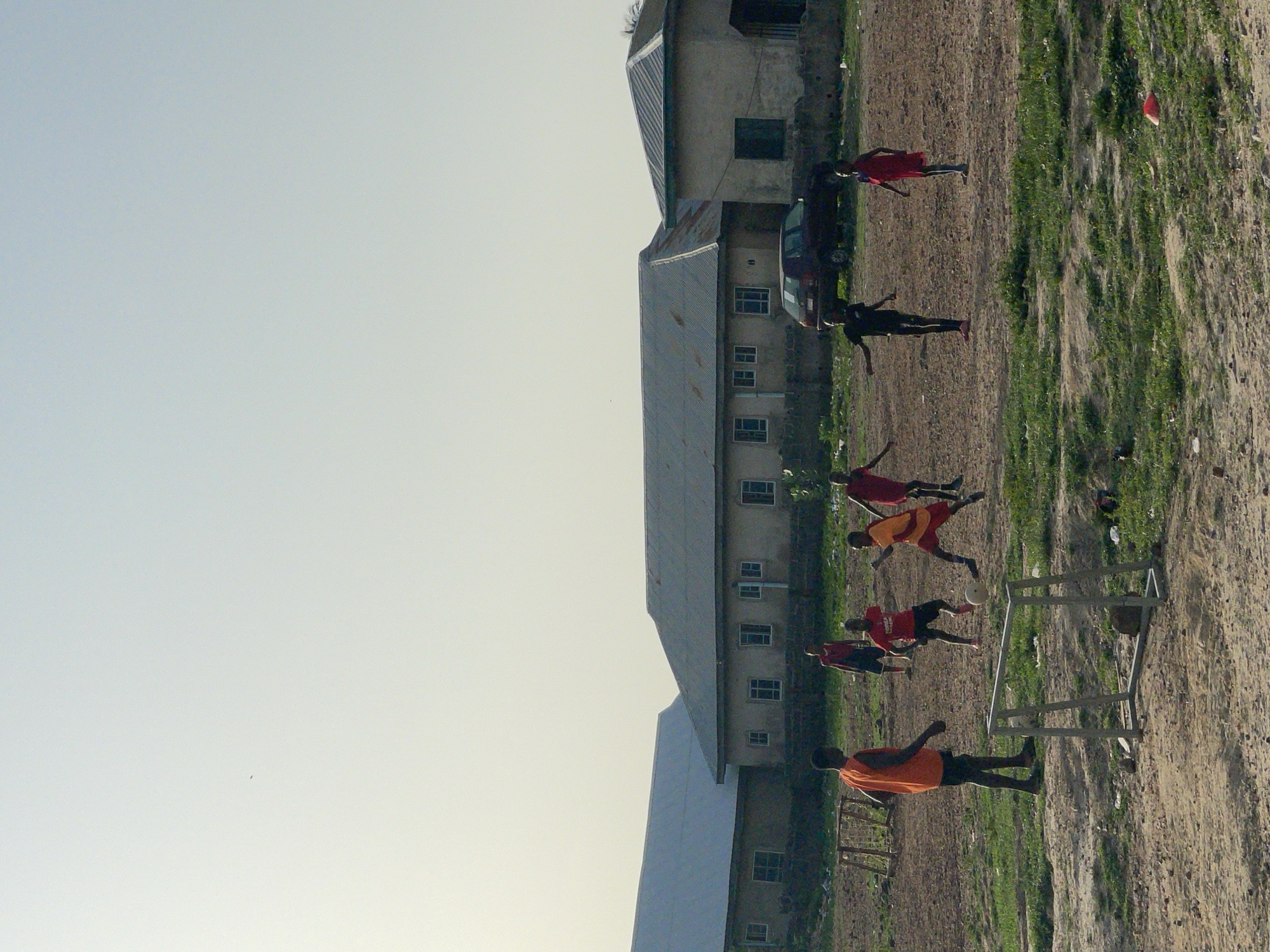 The image shows an outdoor soccer game in Lokoja, Nigeria. Several young people wearing athletic clothing are playing on a field with a soccer goal. In the background, there are buildings, a car, and sparse vegetation. The sky is light-colored and appears to be daytime. The ground is a mix of dirt and grass.