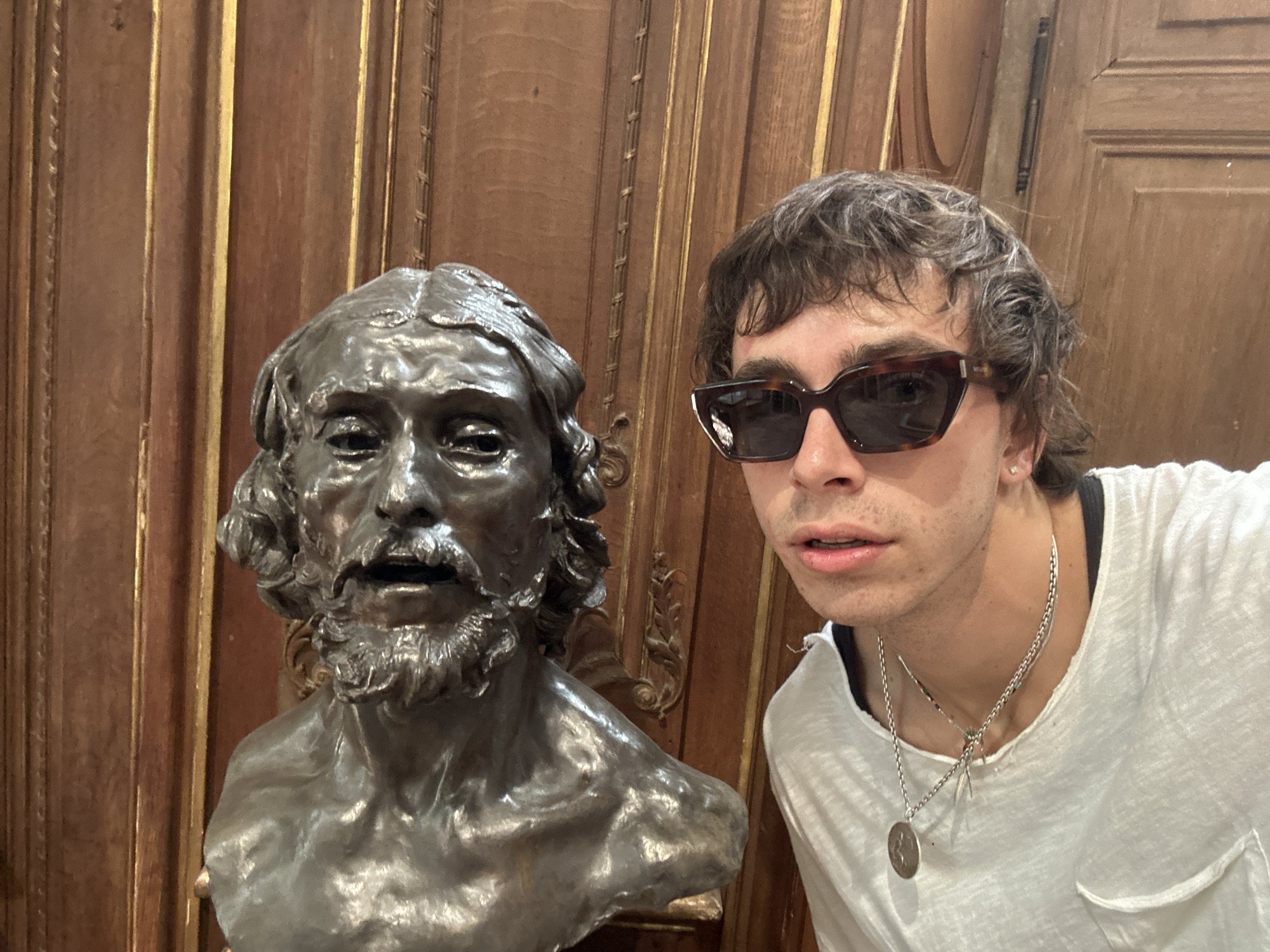 A person with dark hair and sunglasses is taking a selfie next to a bronze bust of a bearded man with curly hair. The person is wearing a white t-shirt and a silver chain with a pendant. They are standing inside a building with ornate wooden paneling on the walls. The photo was taken inside the Palace of Versailles in Paris, France.