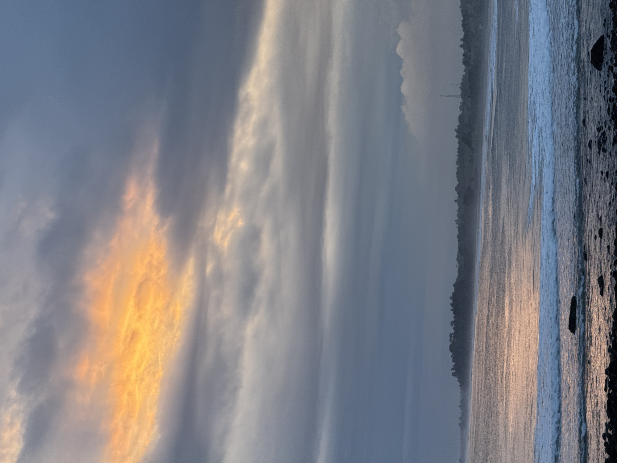 The image shows a beach scene at dusk or dawn in an unknown location. The sky is dominated by gray and white clouds with a vertical streak of orange, indicating the presence of the sun. A dark strip of trees lines the coast, with a thin, tall structure visible through the trees. The beach itself transitions from light-reflecting, wet sand to breaking waves, culminating in a rocky shoreline to the right. No people are visible.